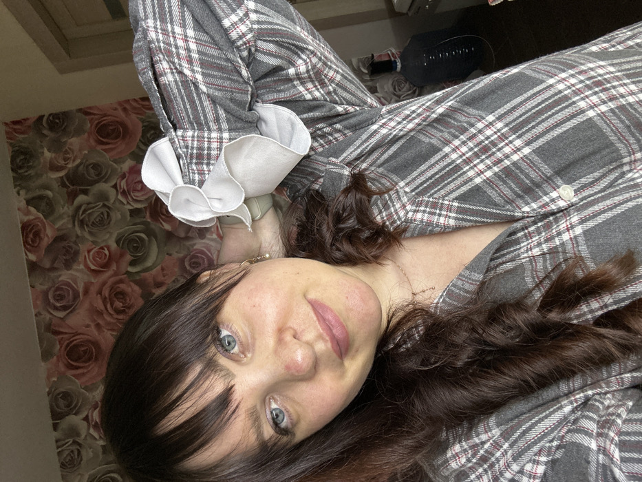 An adult female subject is depicted in a close-up, indoor selfie. She has dark brown, wavy hair with bangs, light blue eyes, and light skin tone. Visible on her face are slight redness on the nose and cheeks, and she is wearing a neutral-toned lip color. She is adorned with a small, light-colored earring in her left ear and a delicate necklace around her neck. The subject is wearing a long-sleeved, grey plaid shirt or pajama top with thin white and red lines, and features ruffled white cuffs. Her left arm is raised, with her hand positioned behind her head/neck area, partially revealing a light-colored wristband. She looks directly at the camera with a slight smile. The background features wallpaper patterned with large, muted roses in shades of pink, red, and olive green. To the right, a segment of a light-colored door frame is visible, along with an out-of-focus dark object resembling a large container with a tube, and a piece of light-colored furniture. The precise location within the city of Noviy, Russia, cannot be ascertained from the visual evidence provided in the image.