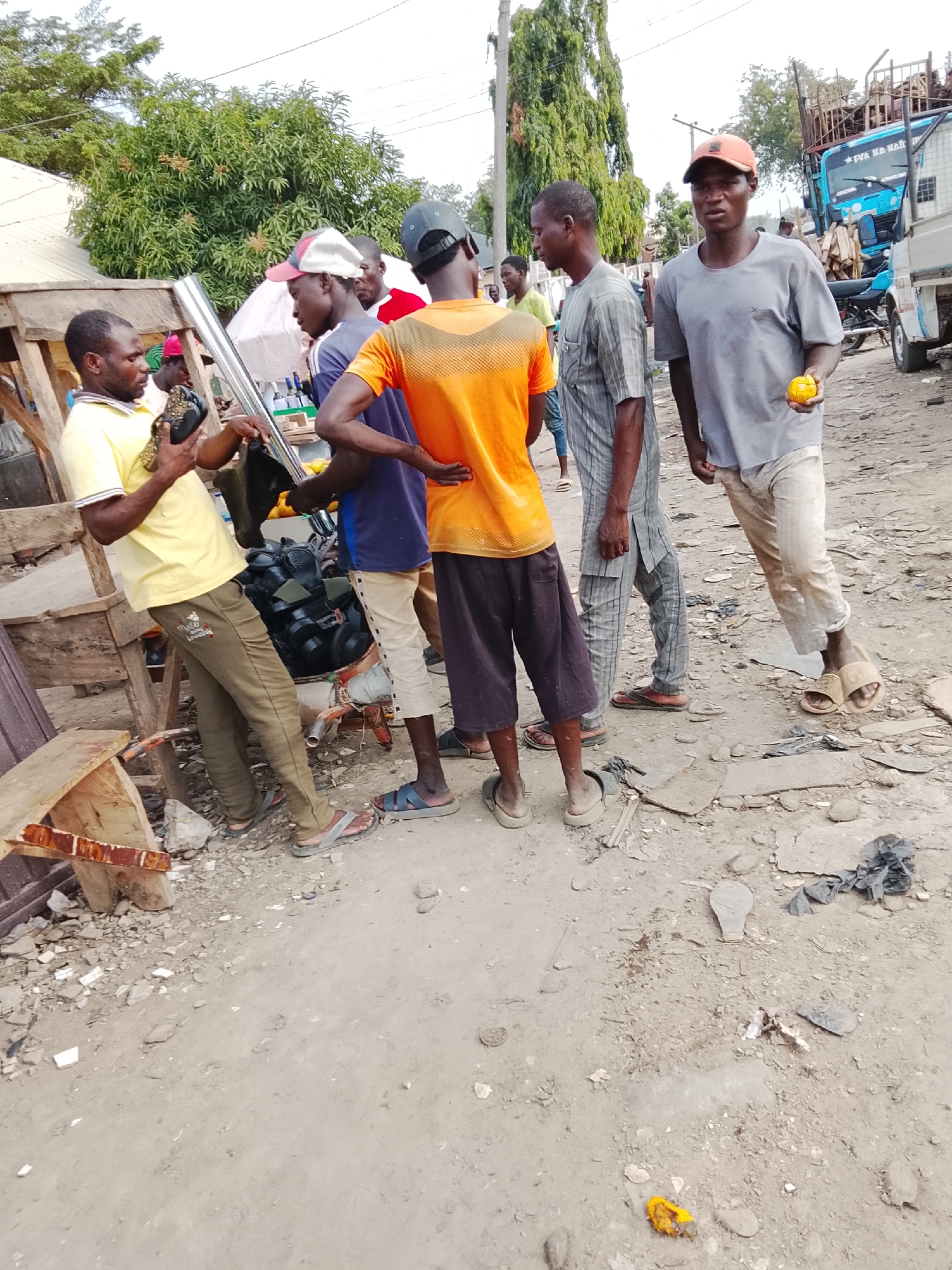 In a dusty area of Abuja, Nigeria, a group of men are gathered. One man, wearing a yellow shirt and brown pants, stands near a makeshift wooden structure, holding up a black object and a long metal tube. Next to him are two men; one in a blue shirt and khaki pants, and the other in an orange shirt and dark shorts. Adjacent to them are two more men: one in a patterned shirt, and the other in a gray shirt, khaki pants, and an orange cap, holding a small orange fruit. In the background, a blue truck is visible. The ground is covered in dirt, rocks, and scattered debris.