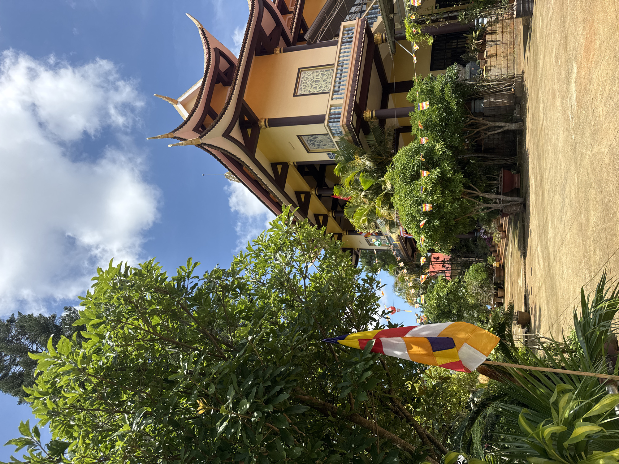 The image captures a vibrant scene in Lạc Dương, Vietnam. A temple, likely a Buddhist temple, is partially visible in the background with a distinctive multi-tiered roof and beige walls accented with dark brown beams and decorative windows. Several colorful Buddhist flags are strung around the temple grounds. In the foreground, lush greenery dominates, including a large tree with thick foliage and various potted plants. A striped Buddhist flag is prominently displayed, its colors of red, white, yellow, and orange catching the sunlight. The sky is a mix of blue and white clouds.
