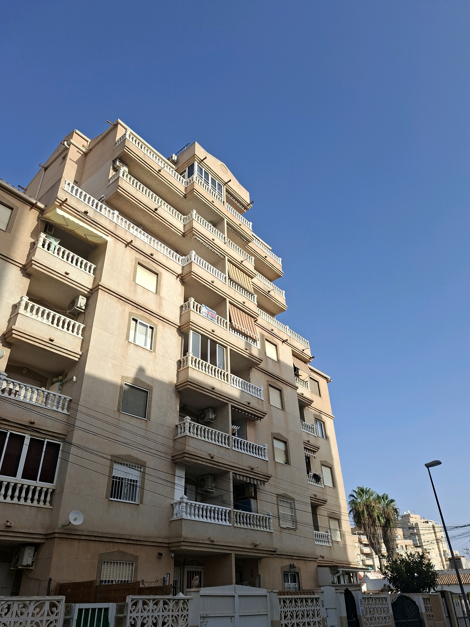 This is a low-angle, full-frame photograph of a beige, multi-story apartment building under a clear blue sky. The building features numerous balconies with white ornate railings and retractable awnings. Several windows are visible, some with blinds. An air conditioning unit is mounted on one of the balconies. A satellite dish is attached to the building's exterior. In the background, another apartment building and a palm tree are visible. A street lamp stands to the right of the frame. The building appears to be located in Torrevieja, Spain, as indicated by the "SE VENDE" (For Sale) sign visible on one of the balconies with a phone number. The ground level shows a decorative fence and a glimpse of a dark green gate.