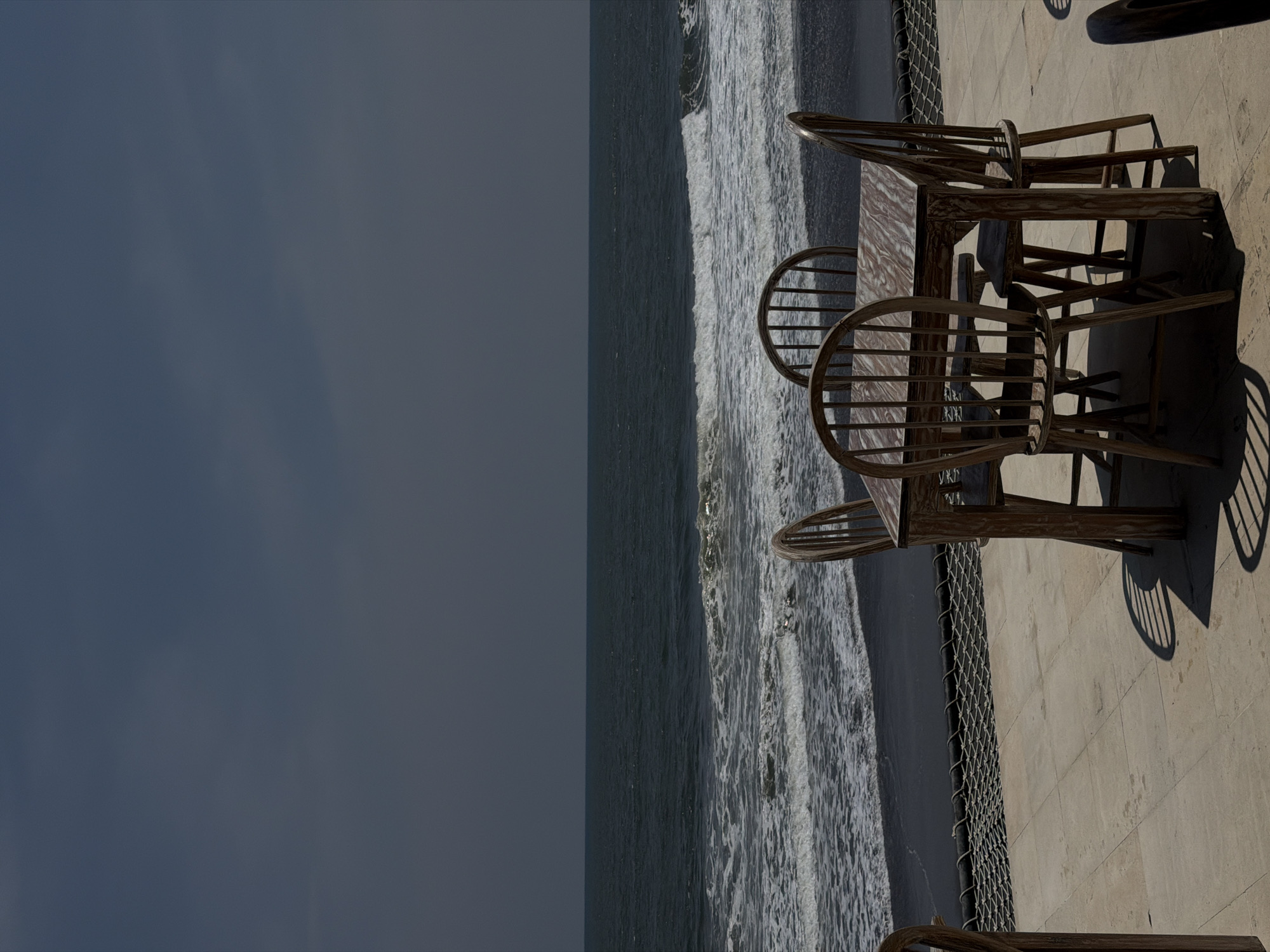 The image displays an outdoor patio scene in Belalang, Indonesia, facing a dark sand beach and ocean under a dark, uniform sky. In the foreground, a light-toned tiled patio floor supports a rectangular wooden table with a patterned surface and four matching wooden Windsor-style chairs, arranged in a dining configuration. Shadows from the table and chairs are cast onto the patio, suggesting direct sunlight. A black mesh net structure is visible along the edge of the patio, bordering the dark sand of the beach. The midground shows ocean waves breaking with white foam near the shore. The background is composed of the open ocean meeting a largely overcast, dark grey sky. No individuals are present in the scene.