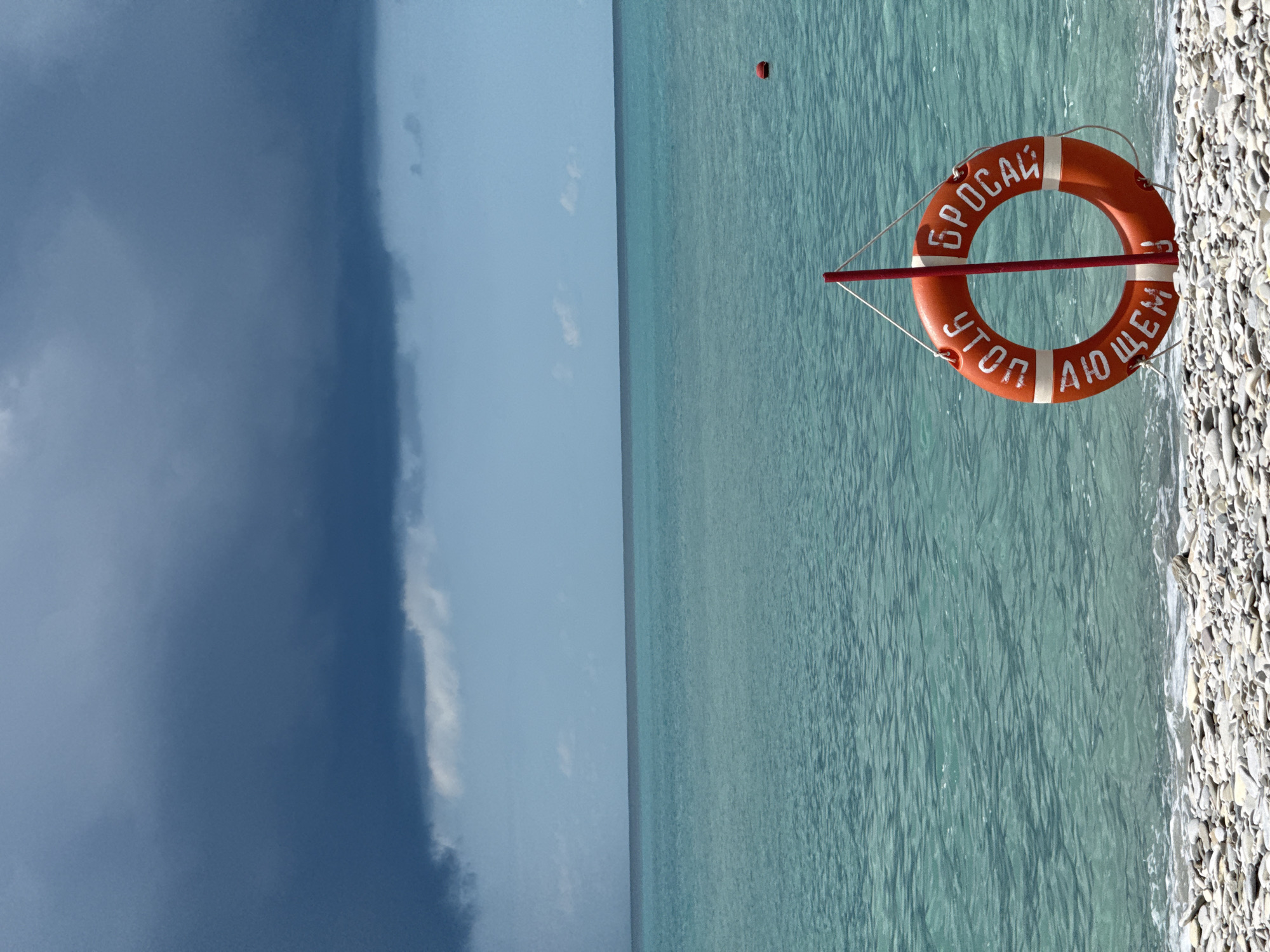 In Nebug, Russia, a lifebuoy is affixed to a red pole on a pebble beach. The lifebuoy is orange with white lettering that reads "БРОСАЙ, КОГДА ТОНЕШЬ" which translates to "THROW WHEN YOU DROWN". The sea is a vibrant turquoise, with gentle waves lapping the shore. Dark clouds loom in the distance, contrasting with the clear blue sky above. A small red buoy floats in the water.