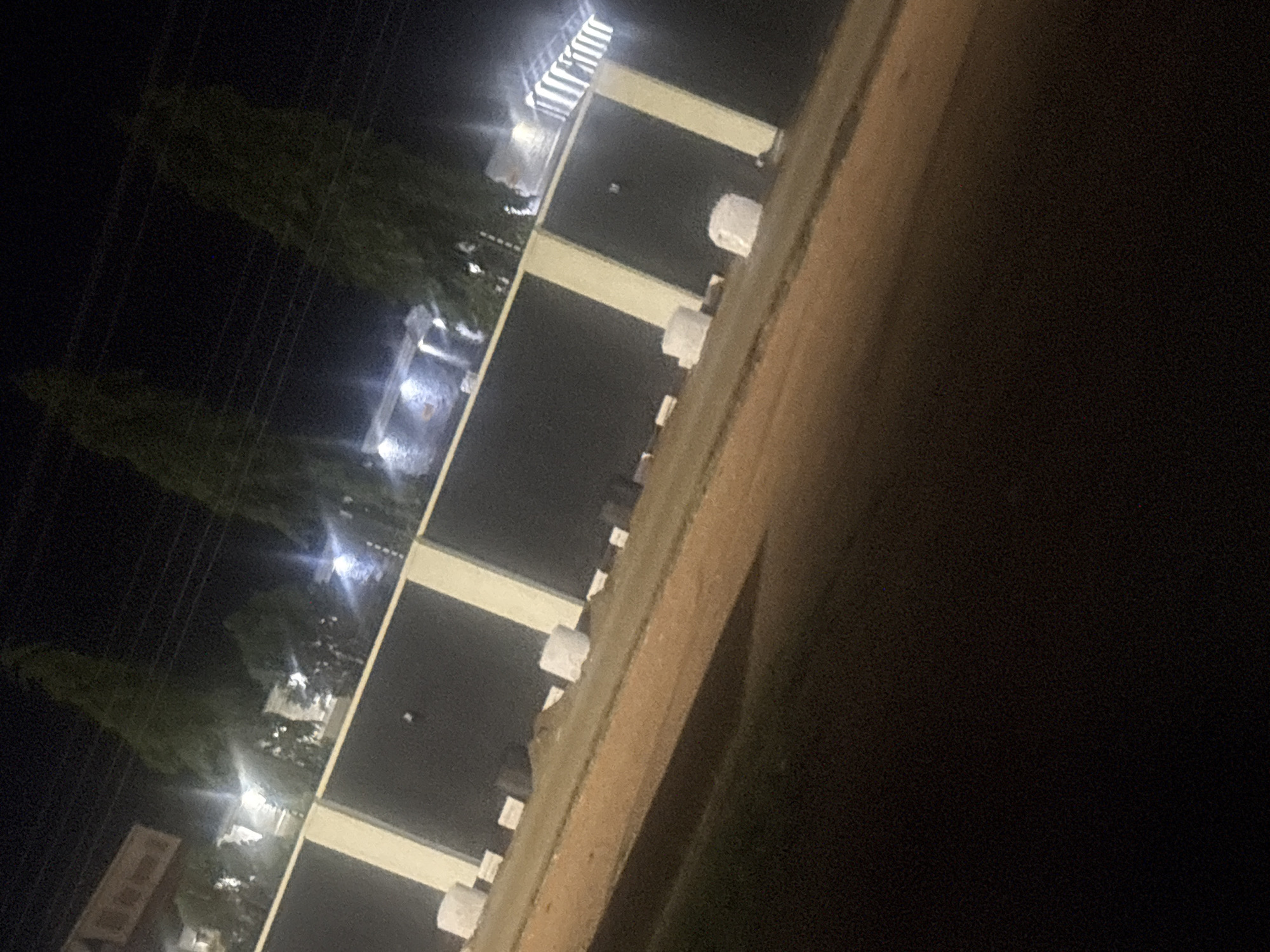 The image shows a nighttime view in Bukuru, Nigeria. A building with multiple stories is visible, featuring evenly spaced vertical supports or pillars connected by horizontal beams. Illumination from interior lights casts a glow on the surrounding areas. Tapered trees line the roadside. Power lines are faintly discernible. A section of a road or elevated surface is apparent in the foreground. The environment is predominantly dark, with limited ambient light.