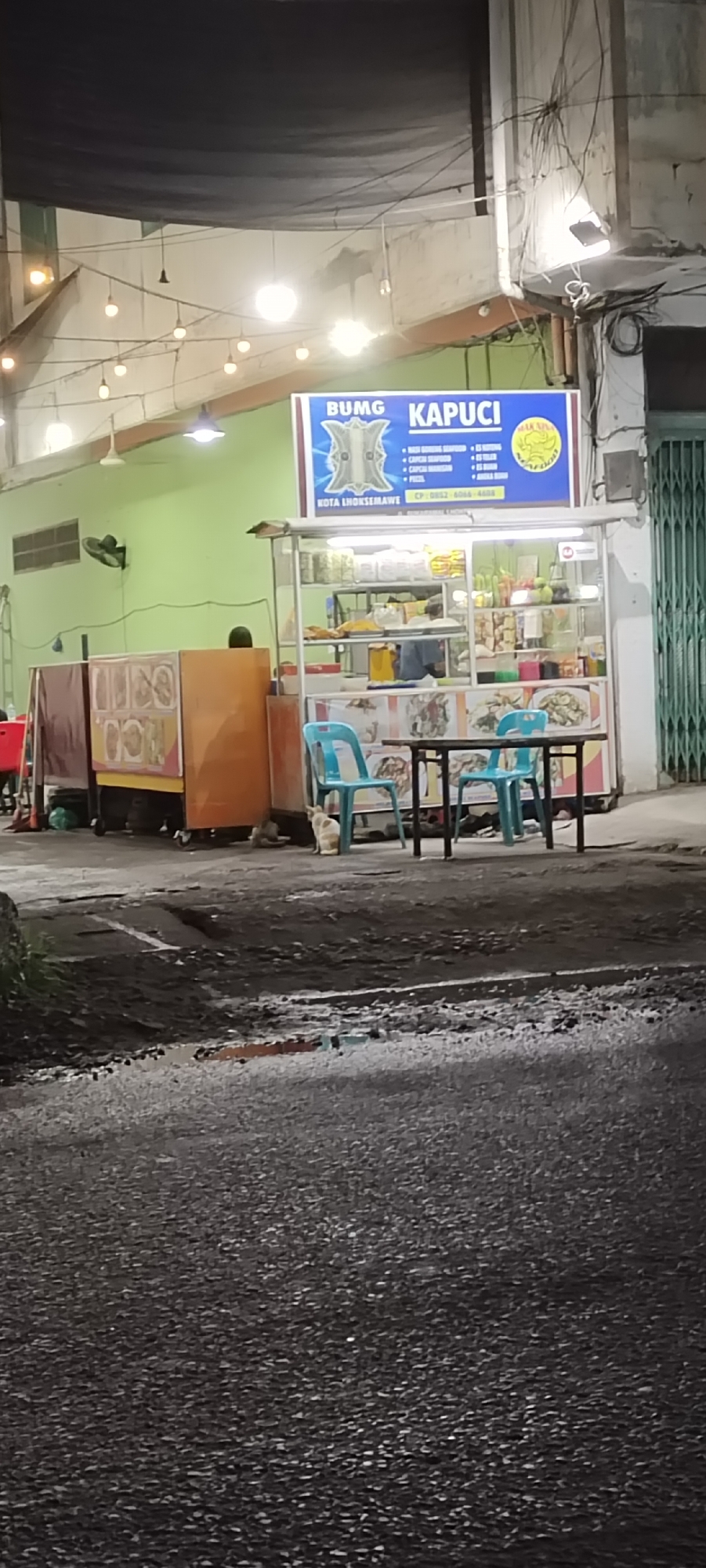 This image captures a nighttime scene of a street food stall in Lhokseumawe, Indonesia. The main subject is a brightly lit food cart, distinguished by a prominent blue sign above it. The sign advertises "BUMG KAPUCI" and lists various menu items including "NASI GORENG SEAFOOD", "CAPCAY SEAFOOD", "CAPCAY MAKANAN", "PIZZA", "ES TELLER", "ES BUAH", and "ANEKA BUAH," along with a "JAKARTA SEAFOOD" logo, the location "KOTA LHOKSEUMAWE," and a contact number.
The food stall, made of white glass and metal, displays food items and condiments. In front of it, two light blue plastic chairs are placed around a dark rectangular table, indicating a small dining area. To the left, other food carts or displays are visible, including one with an orange-brown finish and another with patterned sides, suggesting a cluster of vendors. A person is partially visible behind the main stall, seemingly attending to it. Another figure is faintly seen behind the carts on the left.
The setting appears to be an open-air eatery or a covered section of a building, with light green walls and a dark tarp or awning overhead from which string lights with bare bulbs are strung, casting a warm glow. A bright white security light is mounted on a concrete pillar on the right. A black or dark green metal roll-up door is visible on the far right. A ceiling fan is also mounted on the light green wall.
The foreground is a dark, coarse asphalt road or ground, appearing rough and possibly damp. A light-colored cat with markings sits near the blue chairs, and another small animal, possibly a kitten, is partially obscured nearby. The scene depicts a typical evening street food ambiance, with artificial lights illuminating the area against the dark night sky. The overall mood is casual and inviting, characteristic of urban street life in Indonesia.