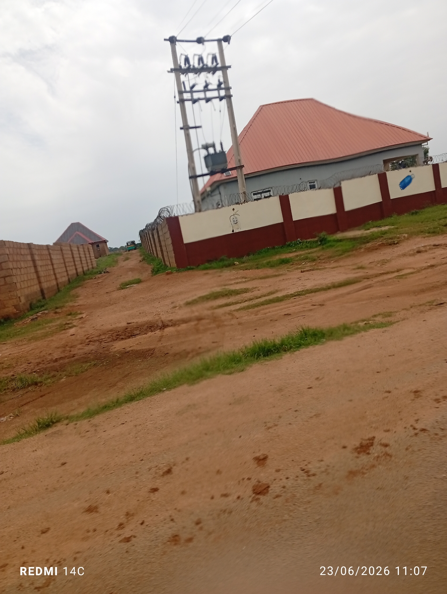 This image depicts an outdoor scene in a semi-arid environment, likely in Bukuru, Nigeria. The foreground is dominated by a dirt road with patches of dry grass. In the mid-ground, a brick wall runs along the left side of the road, and beyond it, another building with a dark roof can be seen. On the right side of the road, a substantial wall with a white and maroon finish, topped with barbed wire, encloses a property. A house with a reddish-orange metal roof and grey walls is visible behind this wall. A tall utility pole with electrical transformers and wires stands prominently near the house. Further down the road, a small, green vehicle, possibly a tricycle or a mini-van, is visible. The sky is overcast, suggesting a cloudy day. The overall impression is one of a developing or partially built-up area.
Visible text in the image includes "REDMI 14C" in the bottom left corner and a date and time stamp "23/06/2026 11:07" in the bottom right corner. The presence of the phone model suggests the image was taken with a Redmi 14C phone. The date stamp indicates the picture was taken on June 23, 2026, at 11:07 AM. There are no people visible in the image. The scene appears to be a typical day with no specific event taking place.