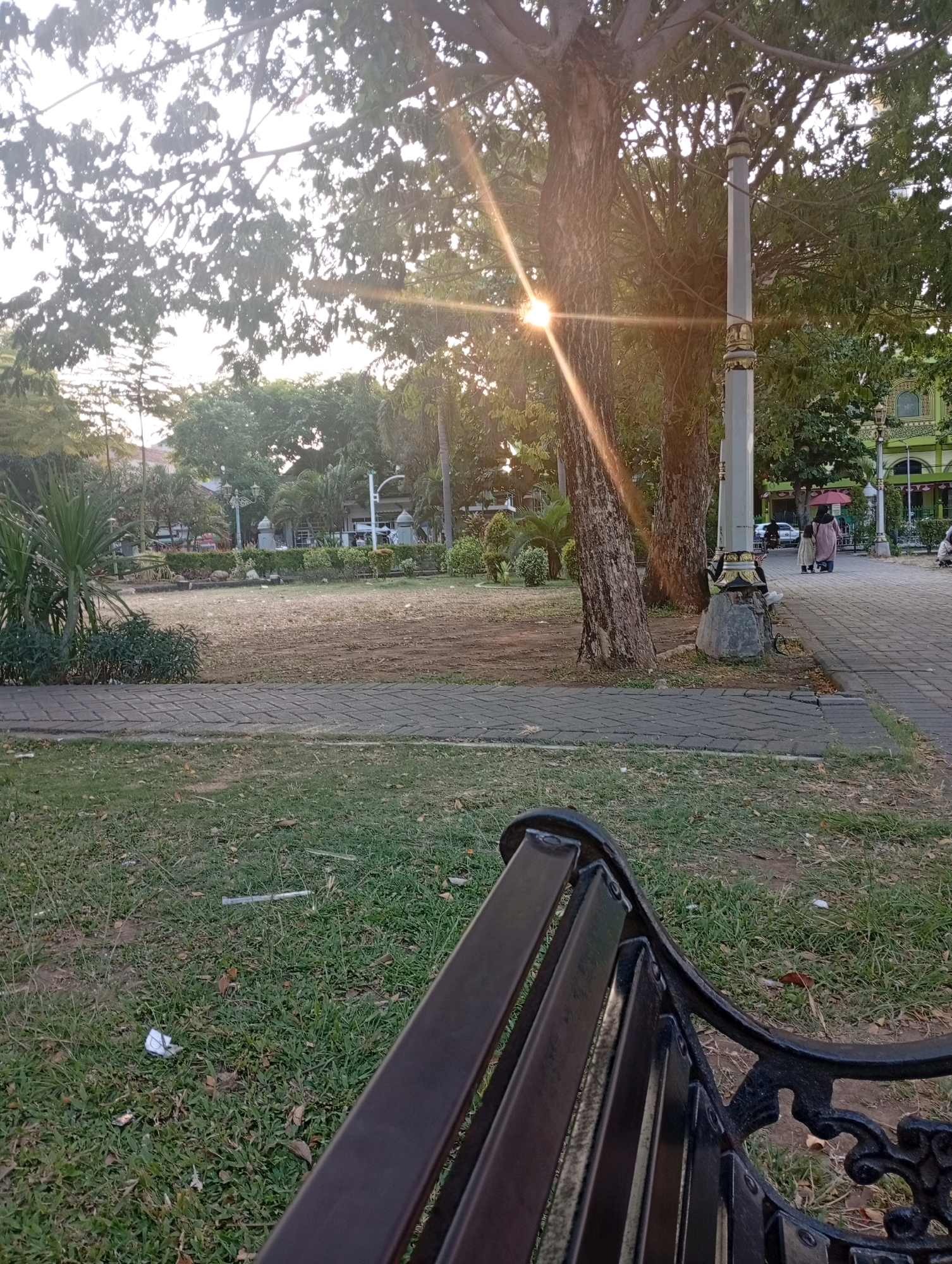 The image depicts a park scene in Bangil, Indonesia, bathed in the warm glow of the setting sun. The foreground is dominated by the back of a dark-colored metal bench. To the left, a patch of green grass is visible, with a few fallen leaves and some scattered litter, including a crumpled white paper and what appears to be a straw. A paved walkway with a brick pattern extends across the middle ground, separating the grassy area from a dirt path. Beyond the path, the park features a dry, patchy lawn with some scattered stones.
Several trees are present, with one large, rough-barked tree positioned prominently in the center-right of the frame. A decorative lamppost with ornate gold detailing stands beside this tree. In the background, there are more trees and some buildings, including a building with green accents and arched windows. A few individuals are visible on the paved walkway; two people, one holding a red umbrella, are walking away from the camera, and another person is seated further down the path. The sun's rays are breaking through the tree canopy, creating a strong lens flare effect in the upper center of the image. The overall atmosphere suggests a quiet, late afternoon in a public park.