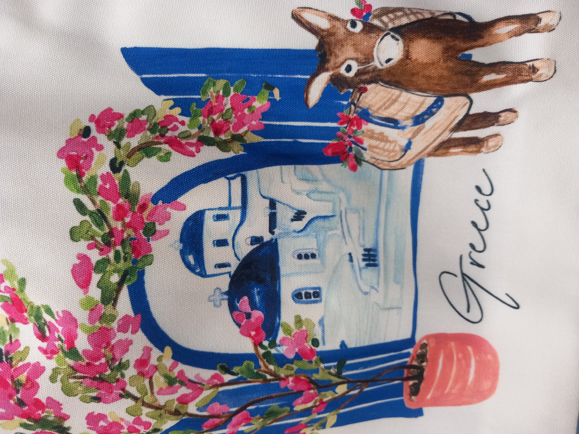 The image is a close-up of a white fabric with a printed illustration. The illustration features a whimsical drawing of a donkey wearing glasses and a straw hat, carrying a basket. The donkey is depicted in a cartoonish style, with a brown body, white eyes with black pupils, and pointed ears. It is adorned with small pink flowers.
To the left of the donkey, a large blue letter "G" is outlined, and within its curve is a sketch of white buildings with blue roofs, reminiscent of Greek architecture. A wreath of pink flowers and green leaves encircles the left side of the "G".
Below the donkey and to the right of the "G" is a handwritten word in cursive: "Greece". Further to the right, a small orange pot contains some dark soil and what appears to be a small plant or branches.
The overall scene seems to be a charming depiction of Greece, possibly a souvenir or decorative item. The illustration is colorful, with prominent blues, pinks, and browns against a white background. The style is lighthearted and illustrative. There are no clear indicators of time of day or weather. The emotions conveyed are cheerful and playful.