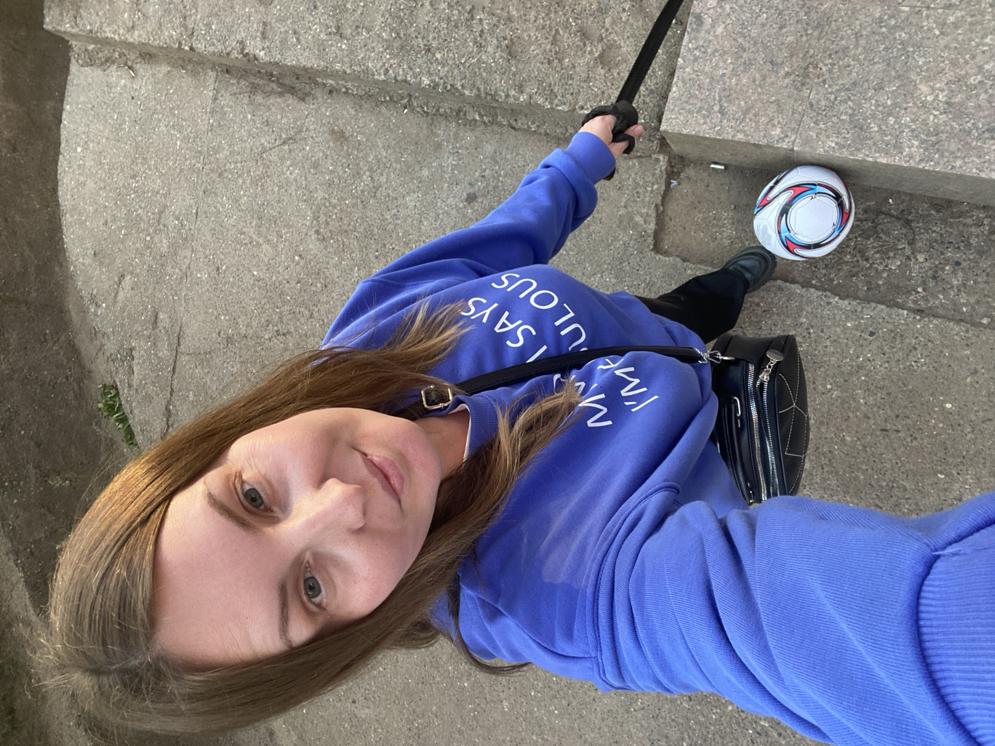 A woman is captured in a selfie-style, slightly angled photograph taken outdoors in Noviy, Russia. She looks directly at the camera with a neutral to pleasant expression, her long brown hair framing her face. She is wearing a periwinkle or lavender-colored long-sleeved sweatshirt. On the chest, white, reversed text is visible, including "SAYS I SA" and "ULOUS," likely forming part of a phrase such as "MY LIFE SAYS FABULOUS." A black shoulder bag with metallic details is slung across her body.
In her right hand, she holds a black pole or stick, which could be a selfie stick or walking aid. The setting consists of grey, textured concrete steps and pavement. To her right, a white football (soccer ball) with distinct black, red, and blue swirling patterns rests on one of the concrete steps. Her left foot, wearing a dark shoe, is partially visible near the ball. The lighting suggests it is daytime. The overall scene depicts a casual outdoor moment.