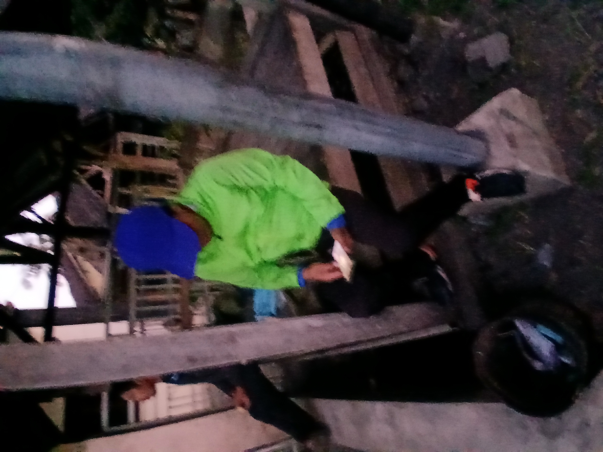 A blurred, low-resolution image captures an outdoor scene at dusk. In the foreground, a large, rough-textured concrete pillar stands to the right of center. To the left of the pillar, a person wearing a bright green long-sleeved shirt and black pants sits on a wooden structure. They are wearing a blue cap and appear to be holding a small, light-colored object in their hands. Partially visible to the left, another person, dressed in dark clothing, is seated. In the bottom left corner, a dark-colored bucket contains some items, appearing to be fish. The background suggests a rustic structure with a thatched or corrugated roof, and possibly a residential building beyond. The overall setting appears to be a village or rural area. The lighting conditions indicate that it is either early morning or late evening.