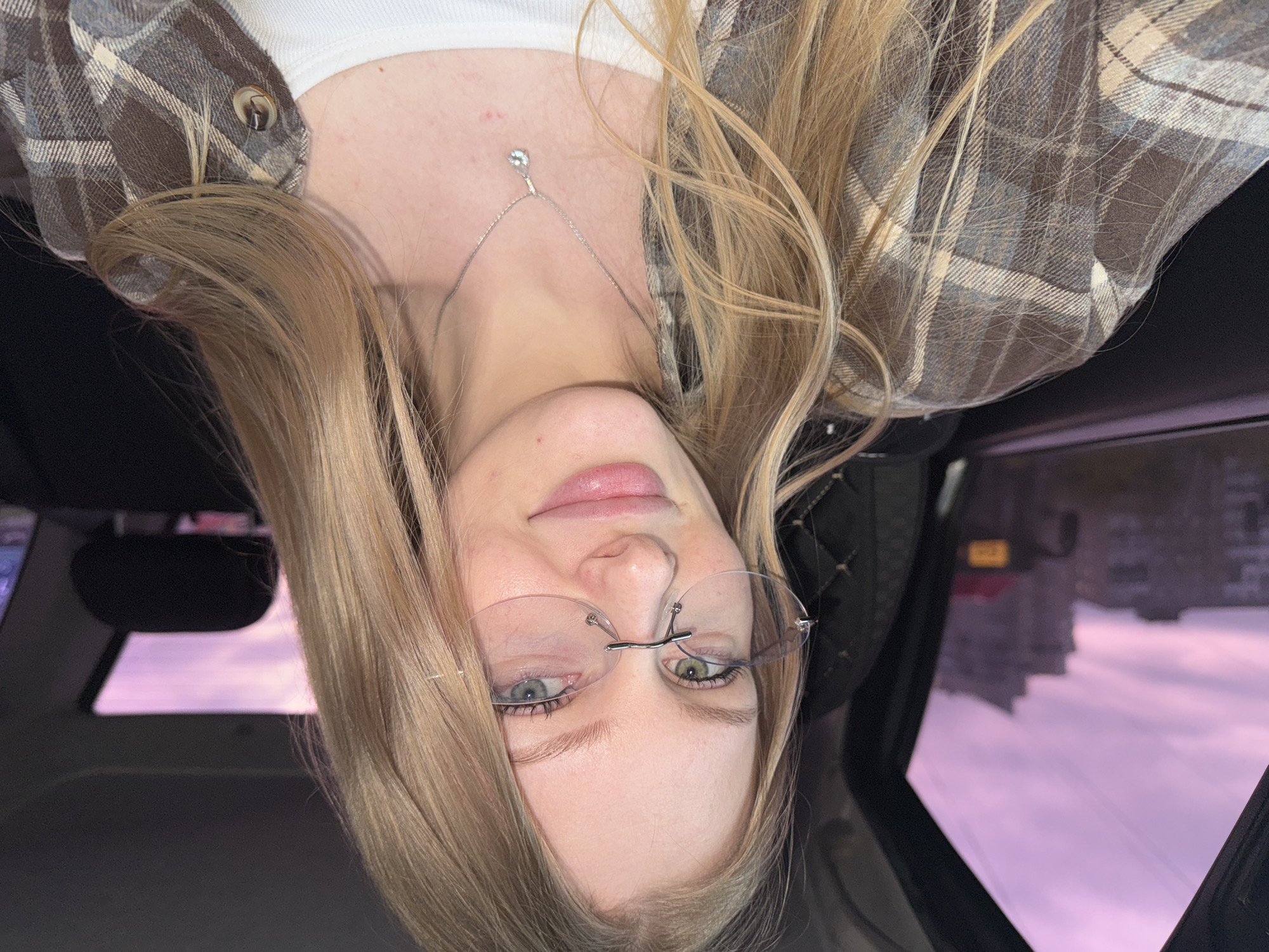 The image shows a woman with blond hair and clear-rimmed glasses, wearing a brown and white plaid shirt over a white top and a silver necklace with a pendant. She is in a car; through the car window one can see a snow-covered landscape with buildings in the distance and a yellowish sign. The sky is pale pink or purple, suggesting dusk or dawn. The reflection in the car window is suggestive of the city of Perm, Russia, due to the architecture and light.