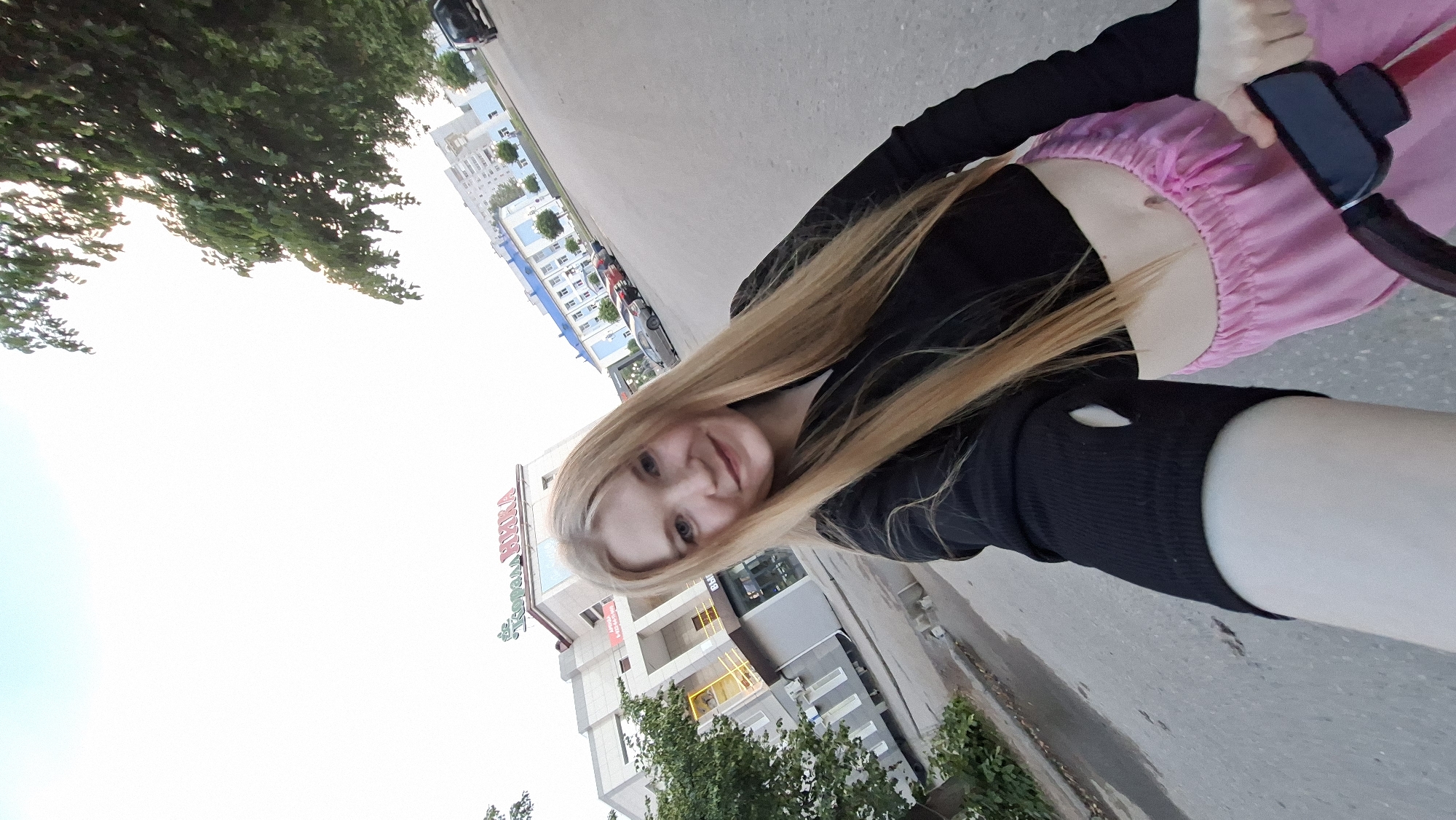 The image shows a young woman with long blonde hair, taking a selfie outdoors in a city. She is wearing a black crop top and pink flowy pants. In the background, there are buildings, cars, and trees, suggesting an urban setting. One building has a sign that appears to be in Russian, with the word "НИКА" (Nika) prominently displayed, and also "АПТЕКА" (Apteka), which means pharmacy. This suggests the location could be near a pharmacy or a business named Nika in Chaikovsky, Russia, a city known for its industrial enterprises and located on the Kama River. The street appears to be asphalt, and there are some puddles on the ground, indicating recent rain or damp conditions. The woman is holding onto what looks like the handle of a stroller or bicycle. The sky is overcast, with some areas of bright white light indicating the sun is likely behind the clouds.