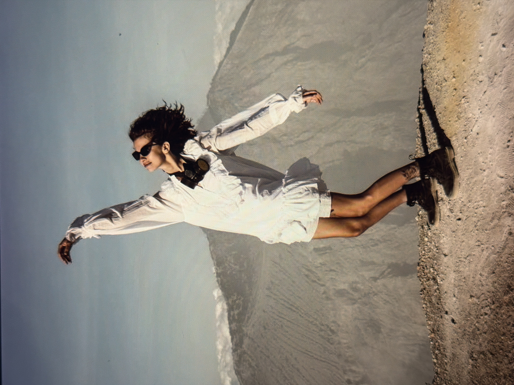 A woman in a white outfit, sunglasses, and boots stands on the edge of a cliff. She is on a rocky cliff edge with a steep drop off to a foggy background. The scene appears to be taken with an effect to make it look like she is flying. The exact location in Đà Nẵng, Vietnam, is not discernible from this image.