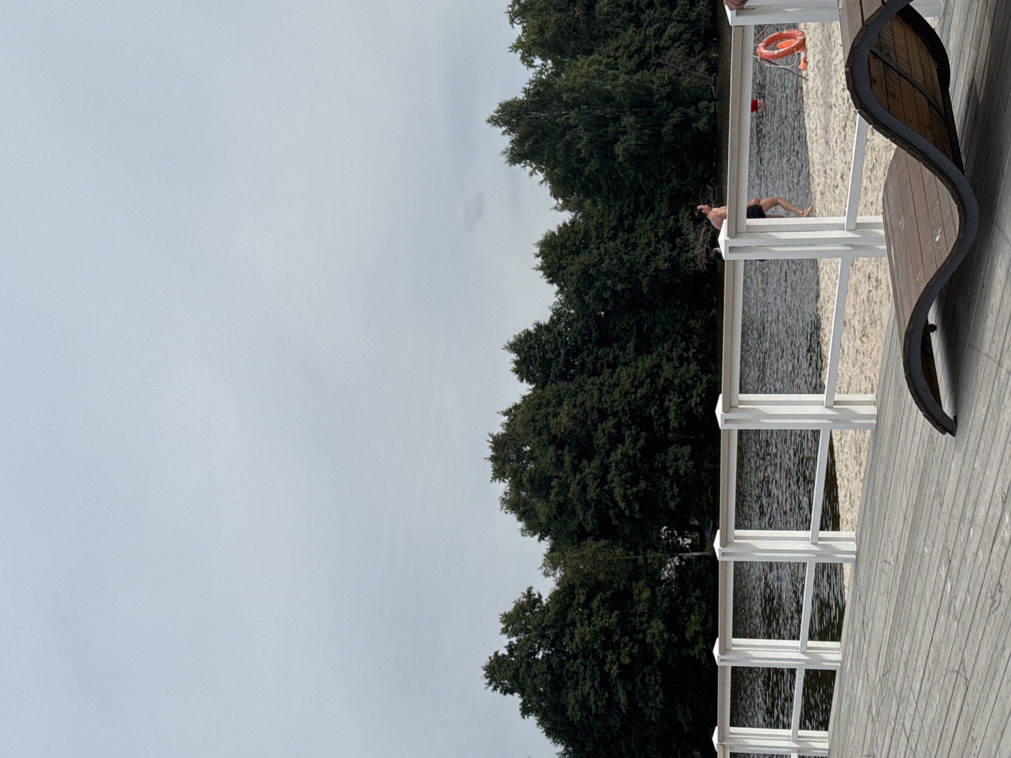 In Sestroretsk, Russia, a person is walking on a sandy beach next to a body of water. The person is wearing black swim trunks. There is a white fence separating the beach from a wooden deck in the foreground. A lounge chair is also visible on the deck. Trees line the opposite shore of the water. The sky is overcast.