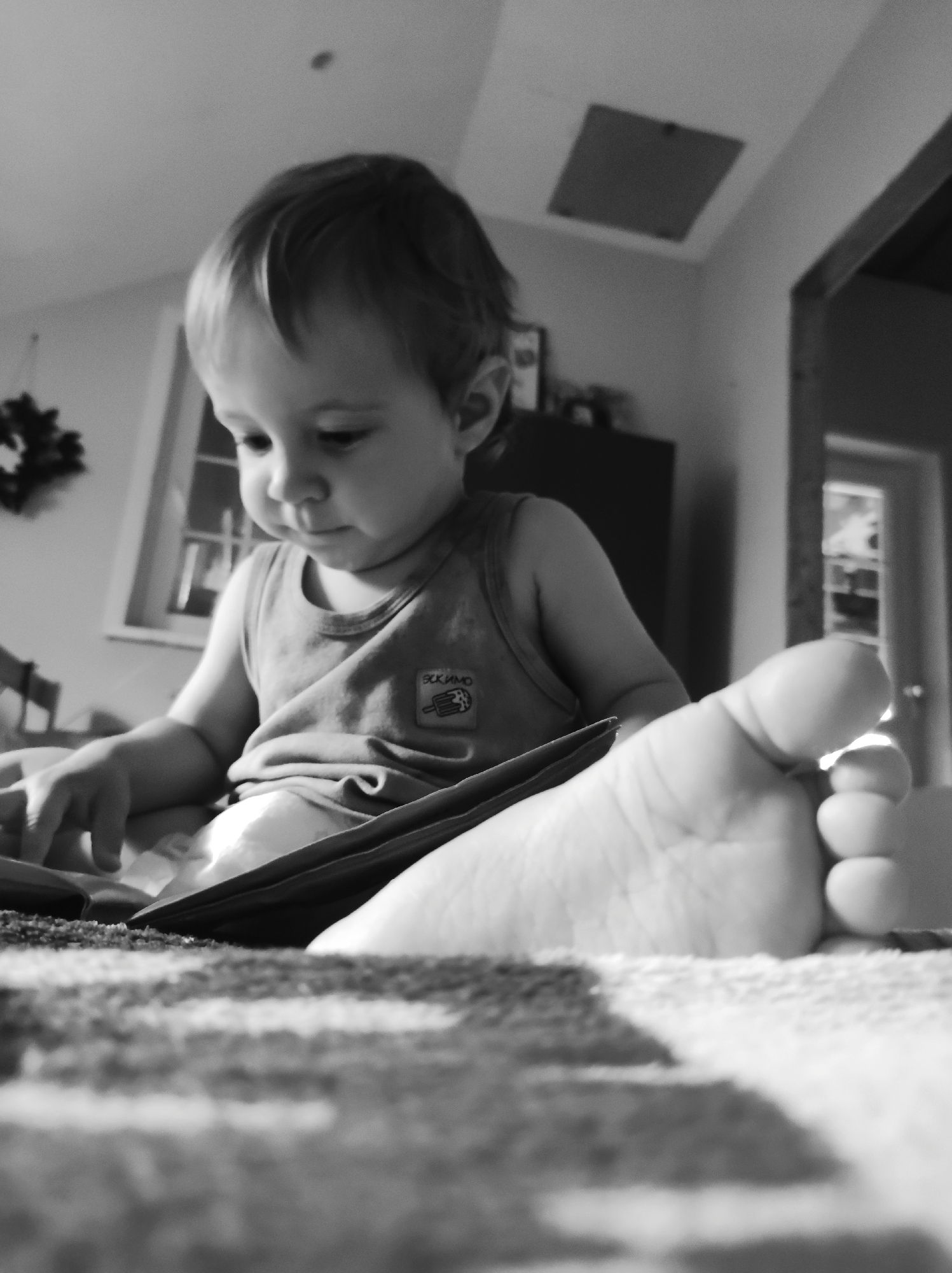 Location: Unknown, Unknown.
A black and white photograph, captured from a low angle, depicts a young child, likely a toddler, seated indoors and looking downward with focused attention. The child wears a light-colored sleeveless undershirt with a dark, rectangular patch on the left chest featuring a lighter graphic, possibly an ice cream cone, and text that appears to be Cyrillic. The child's left hand rests on an unseen object in the lower central frame. In the extreme right foreground, a bare adult foot, with its sole facing the camera, occupies a significant portion of the frame, creating a sense of proximity.
The background shows a domestic interior. To the left, a window with visible panes allows natural light into the space, and a dark, circular decorative item, possibly a wreath, is visible near it. The ceiling above is light-colored and features a rectangular access panel or vent in the upper center-right. To the far right, a dark architectural element, possibly a doorframe or archway, leads into an adjacent room where a door or window with vertical elements can be discerned. The immediate foreground consists of a textured, patterned surface, likely a rug or blanket.
