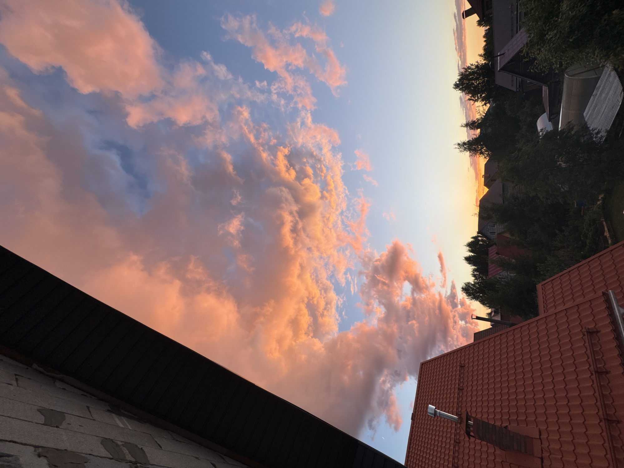 The image depicts a suburban landscape at sunset, characterized by a vibrant sky filled with pink and orange clouds against a clear blue background. In the foreground, a section of a dark, sloped roof is visible on the left, partially obscuring the view of the sky. Below this roof, a red-tiled roof of a house is prominent, with a metal chimney pipe protruding from it. Several other houses with red and dark roofs are scattered throughout the scene, interspersed with lush green trees. The overall impression is of a tranquil evening in a residential area. This scene is consistent with residential areas in the city of Perm, Russia, particularly those with dachas or suburban homes.