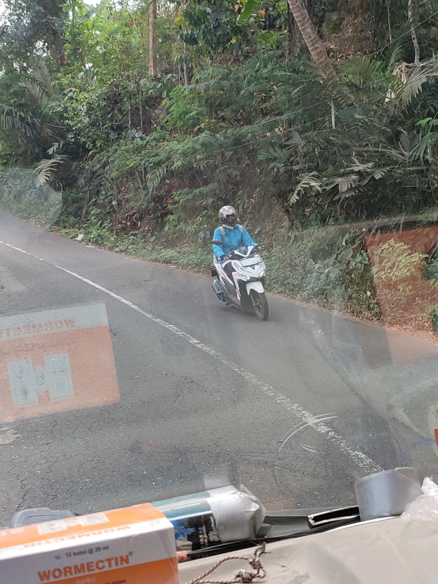 A person wearing a blue shirt and a white helmet rides a white scooter on a paved road in a rural, hilly area with dense green vegetation. The road has a white dashed line. In the foreground, part of a car's interior is visible, with a box labeled "WORMECTIN" in orange and white. A reflection on the car's windshield shows text that appears to say "MORINAG" and some boxes. The scene suggests a journey through a tropical or subtropical landscape.