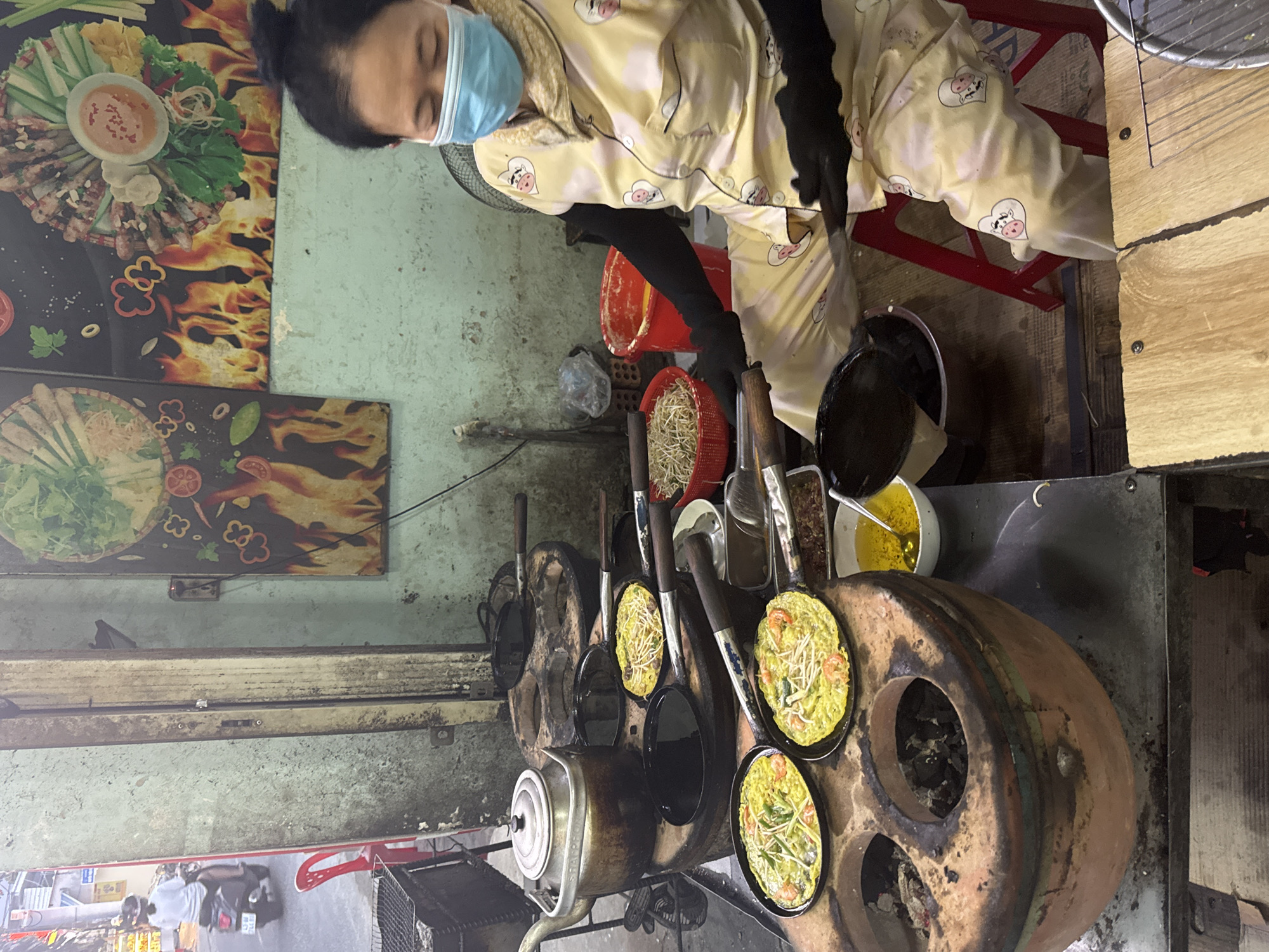The image captures a bustling street food stall in Đà Nẵng, Vietnam, centered around a vendor preparing food. A woman, the primary subject, is shown from the waist up, actively cooking. She wears a light blue surgical mask, a distinctive yellow pajama set with cartoon cow faces inside hearts, and black long-sleeved undergarments or gloves. Her dark hair is pulled back.
The cooking station is a rustic, traditional setup featuring a large, round, multi-holed stove, likely fueled by charcoal. Several small, round pans are placed into these indentations, each holding a small, golden-yellow pancake, probably Bánh Khọt. These pancakes are visibly topped with cooked shrimp and fresh green ingredients, such as bean sprouts or herbs. A metal kettle also rests on a part of the stove.
Adjacent to the cooking area, various ingredients and utensils are arranged. A red plastic basket holds a generous amount of fresh bean sprouts, and a bowl filled with a yellow liquid, possibly cooking oil or batter, sits on the counter. A large black pot is also part of the setup.
The background reveals worn walls with visible signs of age and peeling paint. Two large, framed pictures of colorful prepared food dishes, featuring a fiery design, adorn the wall, acting as visual menus or advertisements. Below the cooking counter, a glimpse of what appears to be a street or alleyway suggests an open-air or semi-open setting typical of street vendors. A red plastic chair is visible under the counter, and a metal fan stands behind the vendor. No discernible text is visible on signs or packaging. The scene depicts a functional and busy food preparation environment.
