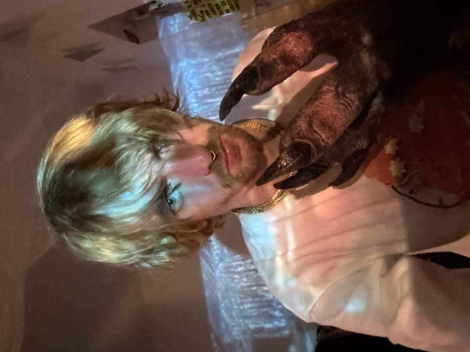 A blond man with a nose ring, light facial hair, and a gold chain necklace poses with a clawed monster hand resting on his shoulder, near a decorated orange object. The scene is lit with an unusual lighting and is rotated for presentation. There is a white sheet or tablecloth in the background.