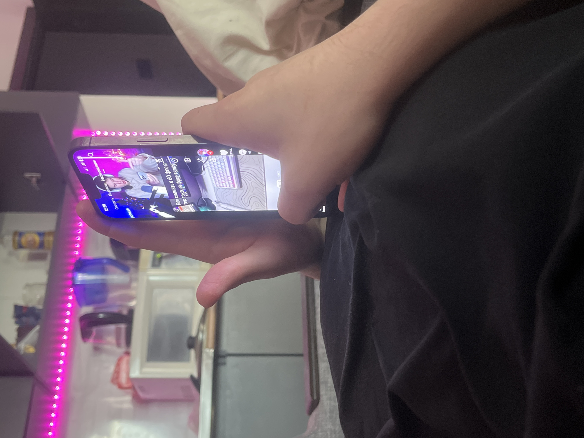 Here is a concise, forensic, matter-of-fact description of the scene:
The image shows a person holding a smartphone in their hands. The phone's screen displays a TikTok video in Russian, featuring a person recording themselves while sitting at a desk with a keyboard and microphone. The TikTok is titled "How to shoot at 60 fps in the front". In the background behind the person holding the phone, there is a kitchen area, including a counter with small appliances and pink LED strip lighting. The environment surrounding the person is blurred. The image is taken indoors. There are no indications of the location being Saint Petersburg, Russia.
