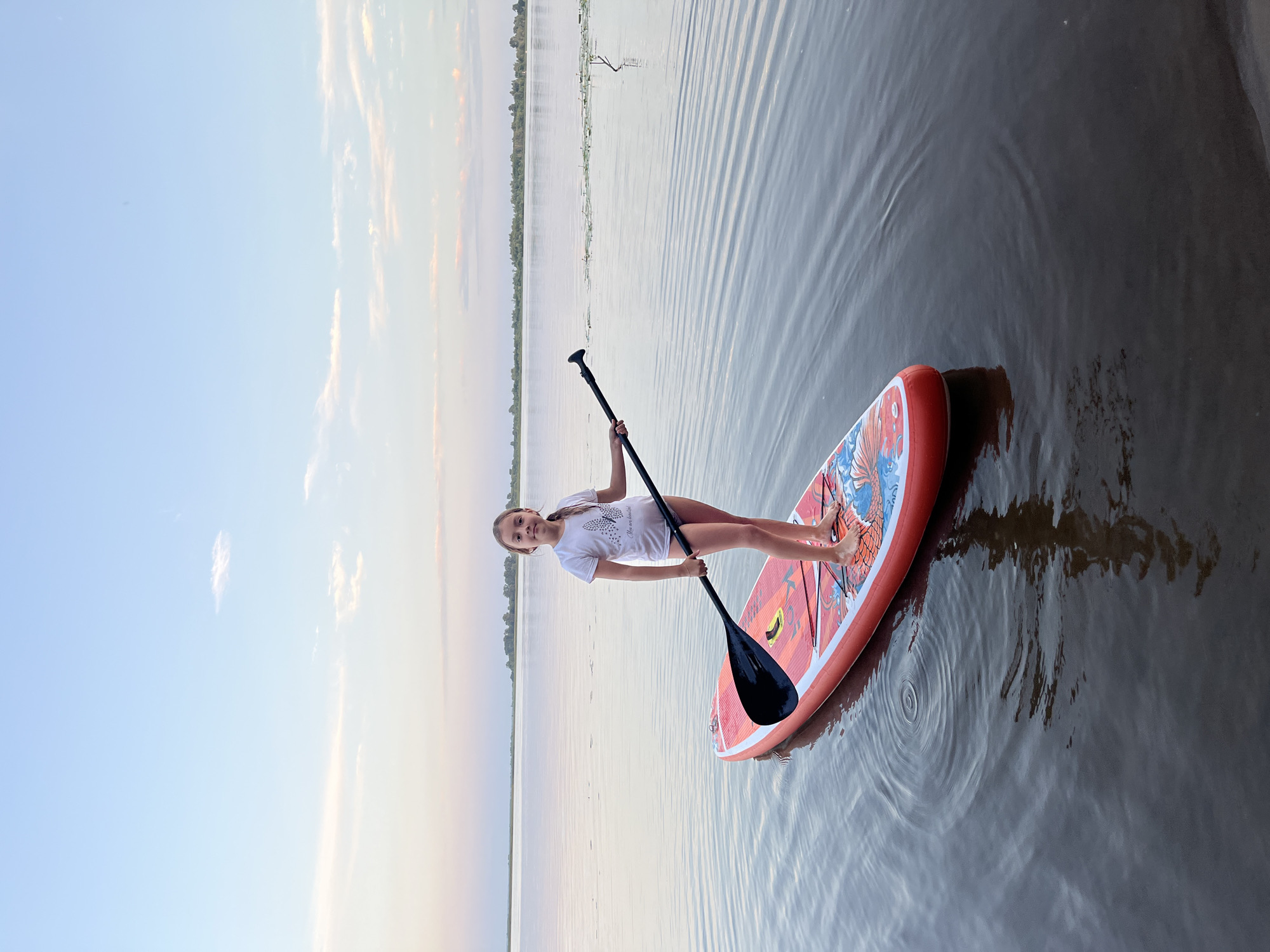 A young girl stands on a red paddleboard in calm water, holding a paddle. She is wearing a white t-shirt with a butterfly design and dark-colored bottoms. The paddleboard has an orange and blue design. The water is still, with gentle ripples emanating from the board. In the background, the sky is a gradient of pale blue and pink, with wispy clouds. The horizon shows a distant shoreline with trees. The scene appears to be at dusk. The location is not specifically identifiable as Ust-Belsk, Russia based solely on the visual information in the image.
