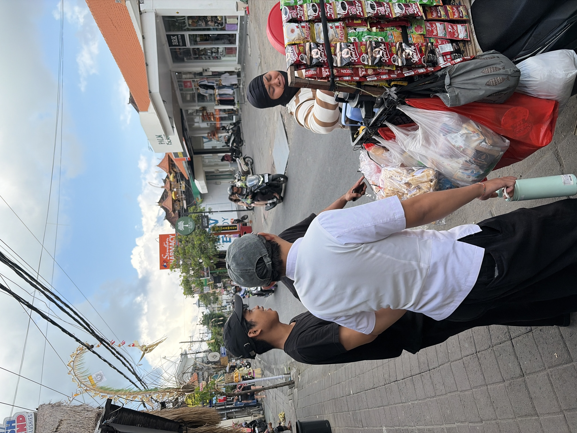 The image shows a street scene in North Kuta, Indonesia, likely during the daytime, as indicated by the bright sky and shadows. Two young men are the primary subjects, with one in the foreground wearing a black t-shirt and a black baseball cap, looking upwards. The other man, slightly behind and to the right, is wearing a white t-shirt and a grey baseball cap, with his arm around the first man's neck. They appear to be interacting casually on a paved sidewalk.
Behind them, a street vendor's cart is laden with various products, including packets of coffee, snacks, and other packaged goods. A woman in a headscarf and striped top is leaning against the cart, her eyes closed, possibly resting or asleep. The cart itself is overflowing with bags of merchandise, some red and white plastic bags holding more items.
The background reveals a bustling street with shops and storefronts. Visible text includes "Semeton PHARMACY" and "BRK COLLECTION," suggesting commercial activity. Several motorcycles are parked or in motion, common in this region. Decorative elements, possibly celebratory kites or banners made of natural materials, are suspended from overhead wires, adding a cultural touch to the scene. The overall atmosphere suggests a lively, everyday street environment in a tropical location.