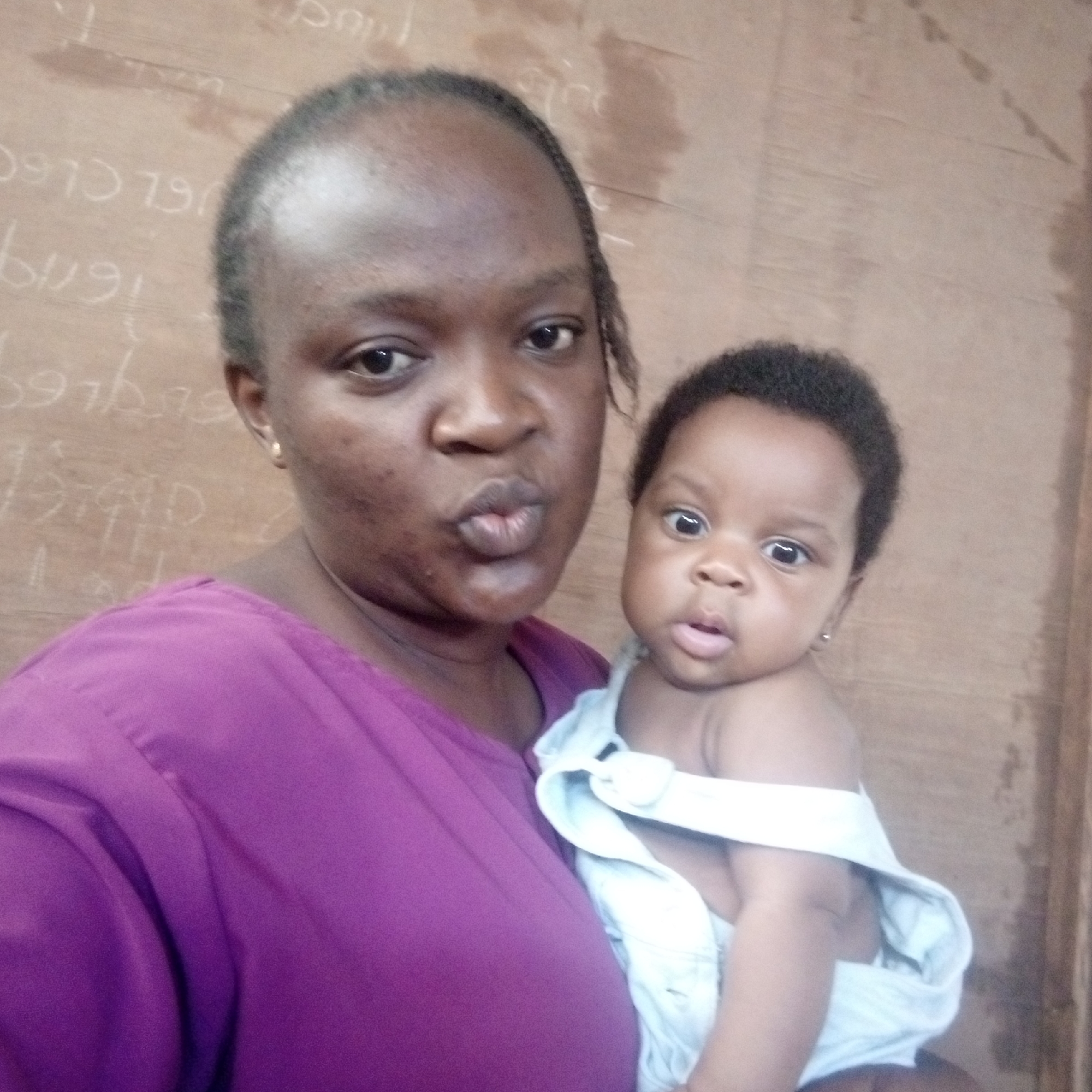A woman is holding a baby in her arms. The woman has dark hair and skin and is wearing a purple shirt. The baby has dark hair and skin and is wearing a light-colored garment. They are inside a room in Ukwu-Abwa, Nigeria, with a wooden wall visible behind them. Writing can be seen on the wall. The woman is puckering her lips. The baby is looking at the camera.
