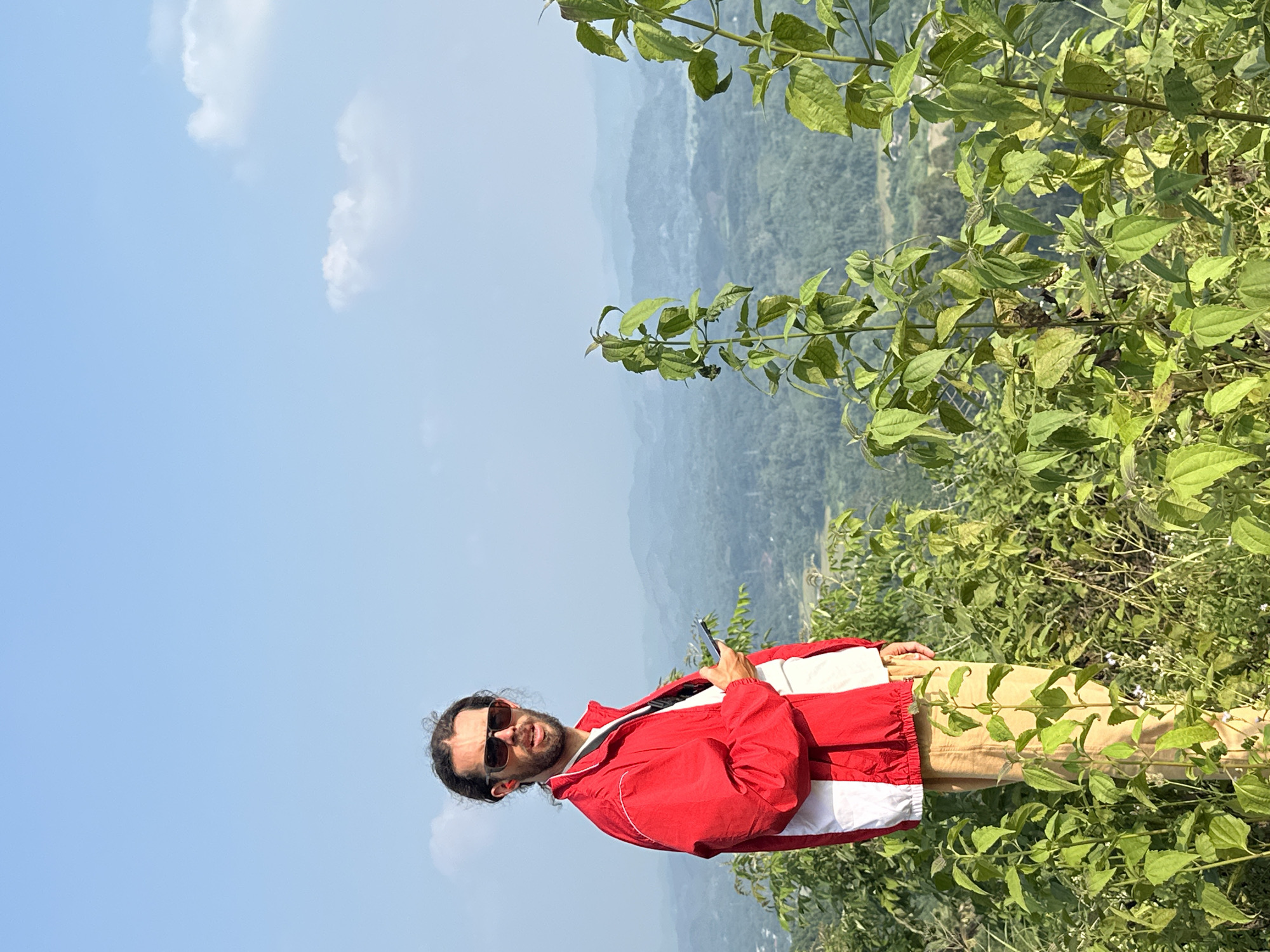 The image, originally rotated 90 degrees clockwise, features a man standing on a scenic hillside under a bright, clear blue sky with sparse white clouds. The setting appears to be an elevated viewpoint in Tân Trịnh Commune, Vietnam, overlooking a vast landscape of green mountains and valleys that fade into atmospheric haze in the distance.
The subject is a man with dark, shoulder-length hair and a beard, wearing sunglasses. He is dressed in a vibrant red jacket with white sections on the sides and sleeves, and light brown or khaki-colored pants. He is holding a dark smartphone in his right hand and looking towards the viewer with a slight smile, suggesting he is posing for the photo.
Lush green foliage and small white flowers are prominent in the foreground and midground, framing the man and the expansive view. The bright daylight indicates it is likely midday. There is no visible text in the image.