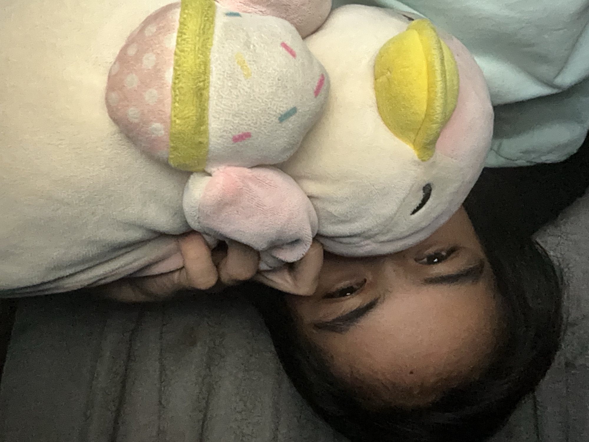 A young individual with dark hair is partially hidden behind a plush toy, peeking out with one eye visible. The person is lying down, with their hand resting on the toy. The plush toy appears to be a pastel-colored character, possibly a bird, with a yellow beak and pink accents. The background is dark and indistinct, suggesting an indoor setting, possibly a bedroom. The overall impression is one of a casual, relaxed moment.
The image does not contain any information that would allow for the identification of a specific location within Davao City, Philippines.