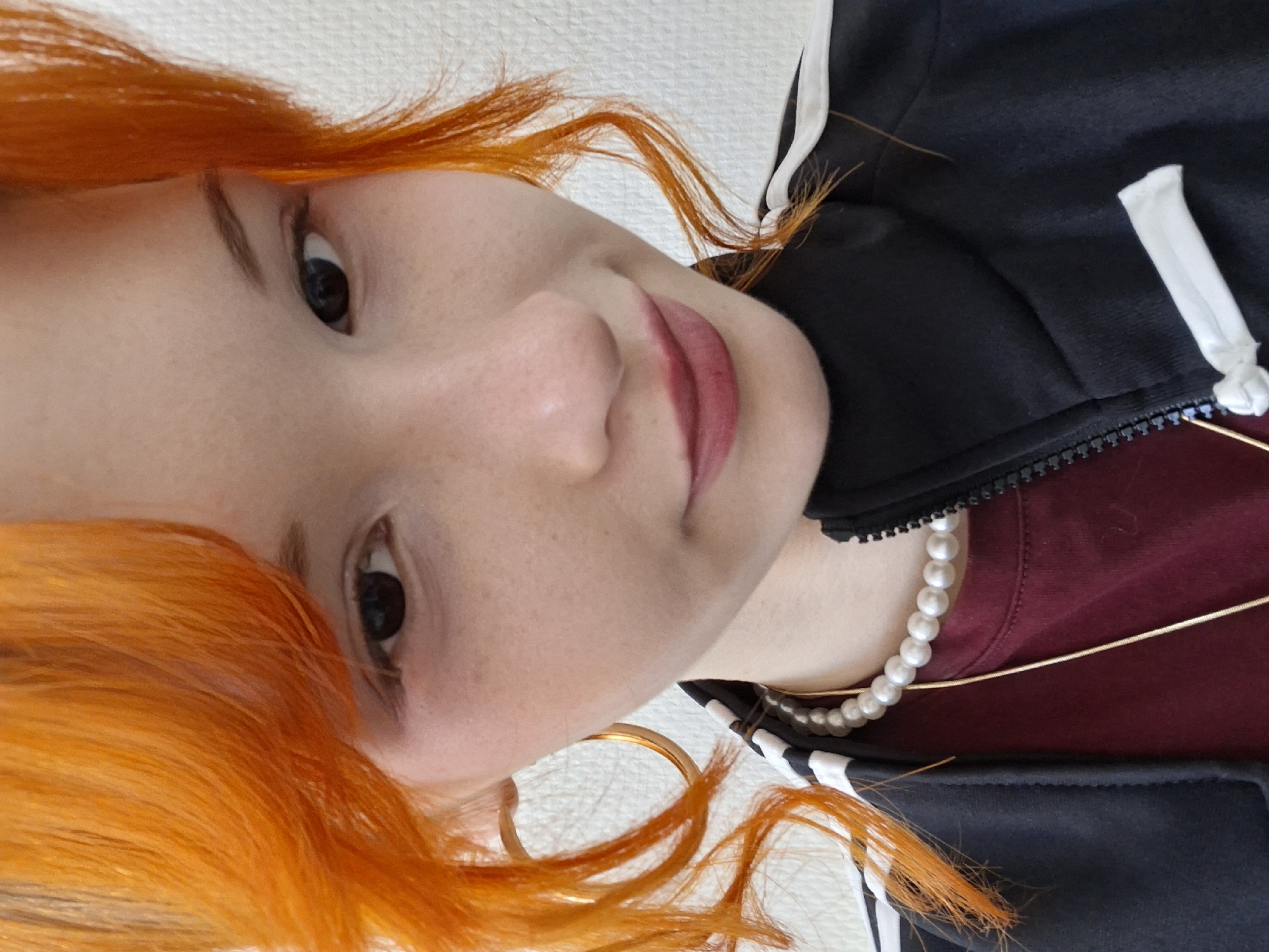 A portrait of a woman with red hair, wearing a black jacket with white stripes, a pearl necklace, and a gold chain. The background is a white, textured wall. The location is Chaikovsky, Russia.