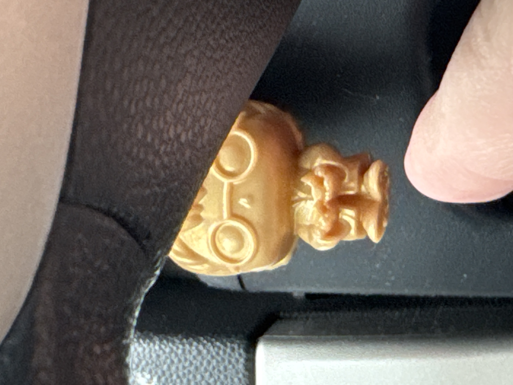 A close-up image shows a small, gold-colored figurine of a person with glasses lying on a dark gray surface. The figurine is partially obscured by what appears to be black fabric on the left and a finger on the right. The environment appears to be indoors, but there are no other details to suggest a specific location, especially within Sofia, Bulgaria.