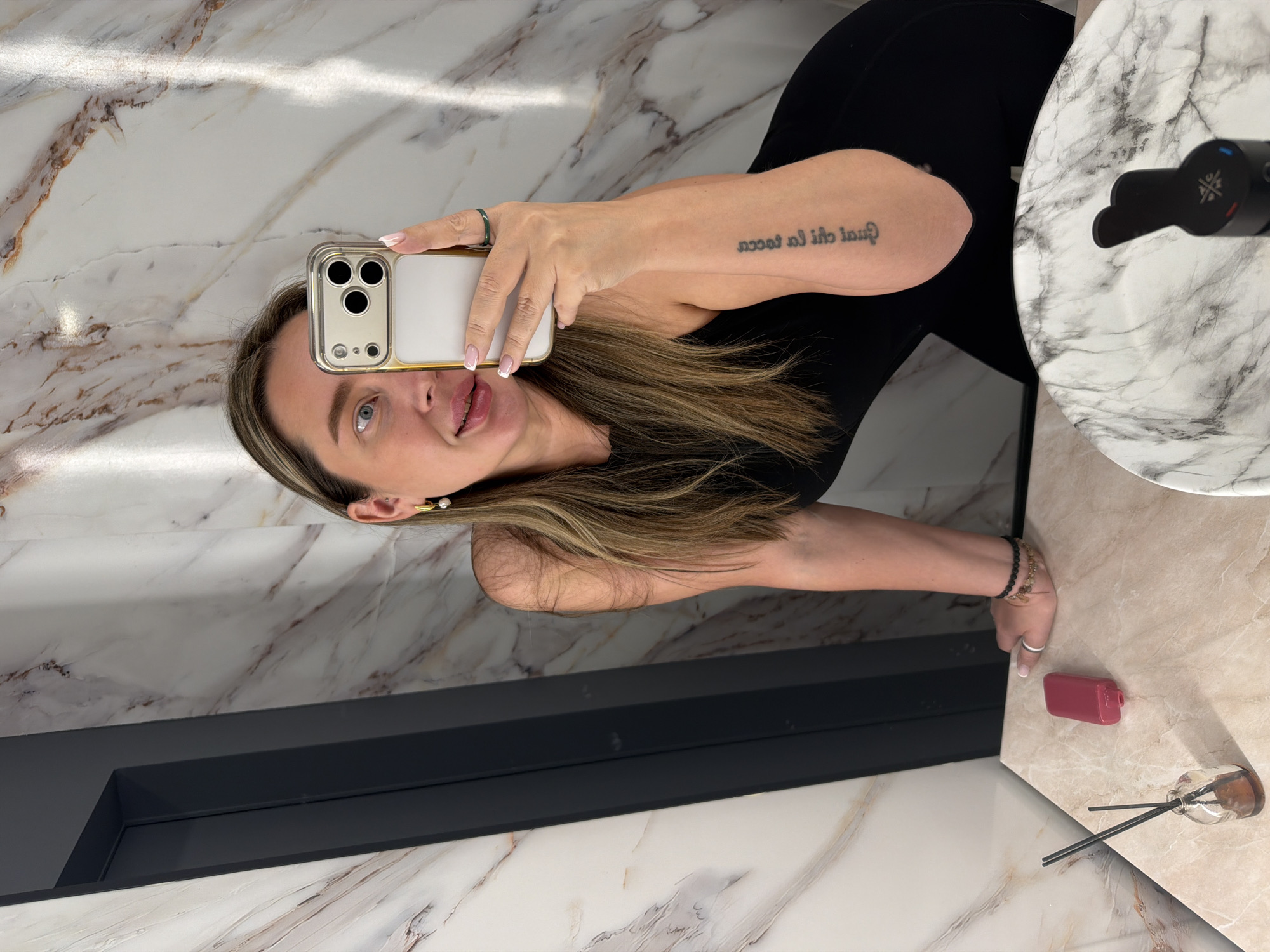 A woman is taking a selfie in a bathroom. She is wearing a black tank top. Her hair is blonde with brown roots. She is holding a white iPhone with a clear case. She has a green ring on her finger. There is a tattoo on her arm. The bathroom has marble walls and a marble countertop. There is a black sink and a black faucet. There is a red object and a glass bottle with black sticks on the countertop. The location is most likely not Moscow, Russia based on the appearance of the bathroom, which doesn't match typical bathrooms in Moscow.
