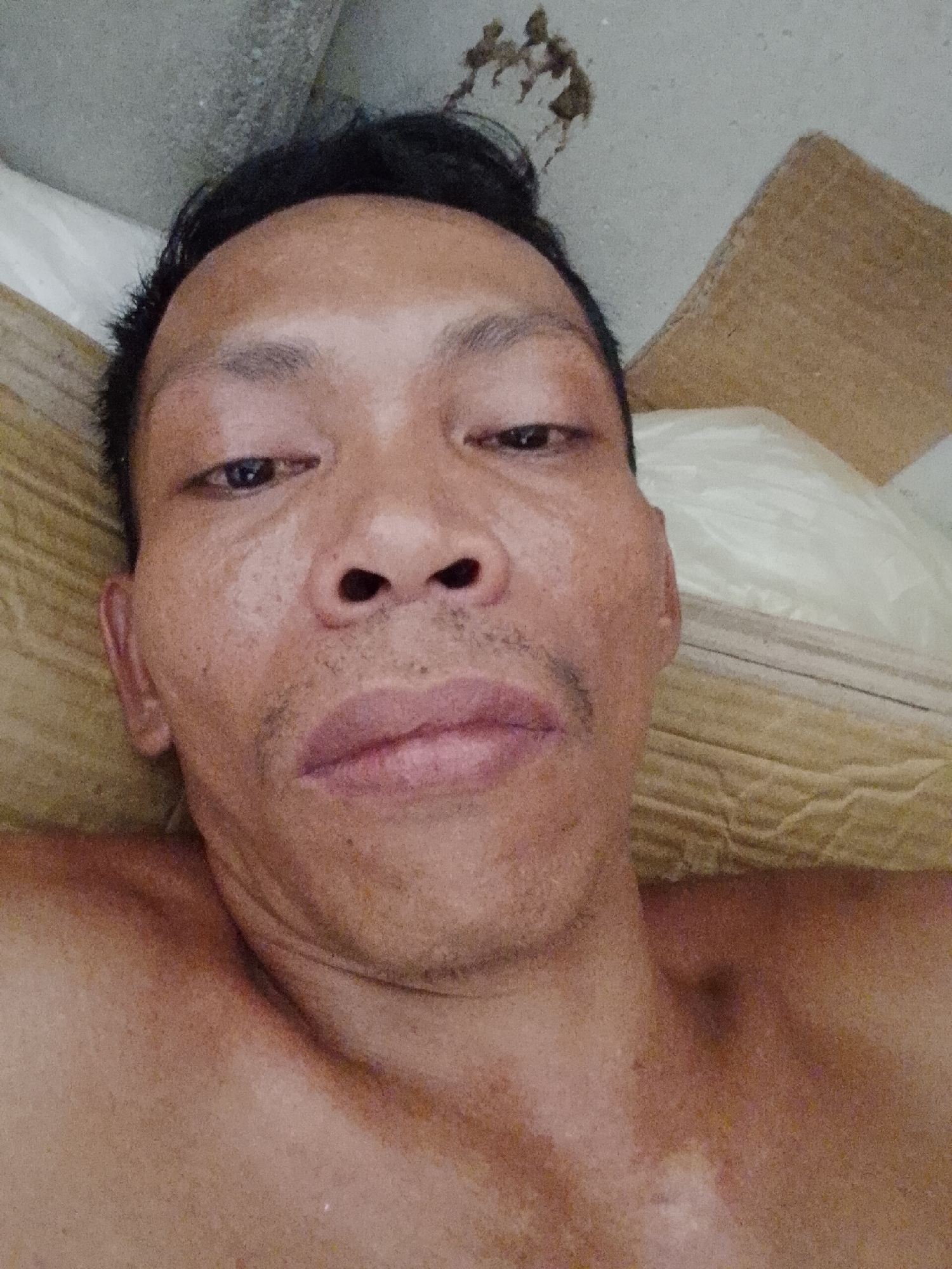 A close-up selfie photograph shows a man with dark hair and tanned skin. He is lying down, possibly on a pillow or bedding, with a piece of cardboard visible behind him. The background includes a gray concrete or cement surface with some discoloration. The photograph was taken at Bukit Ketok, Indonesia.