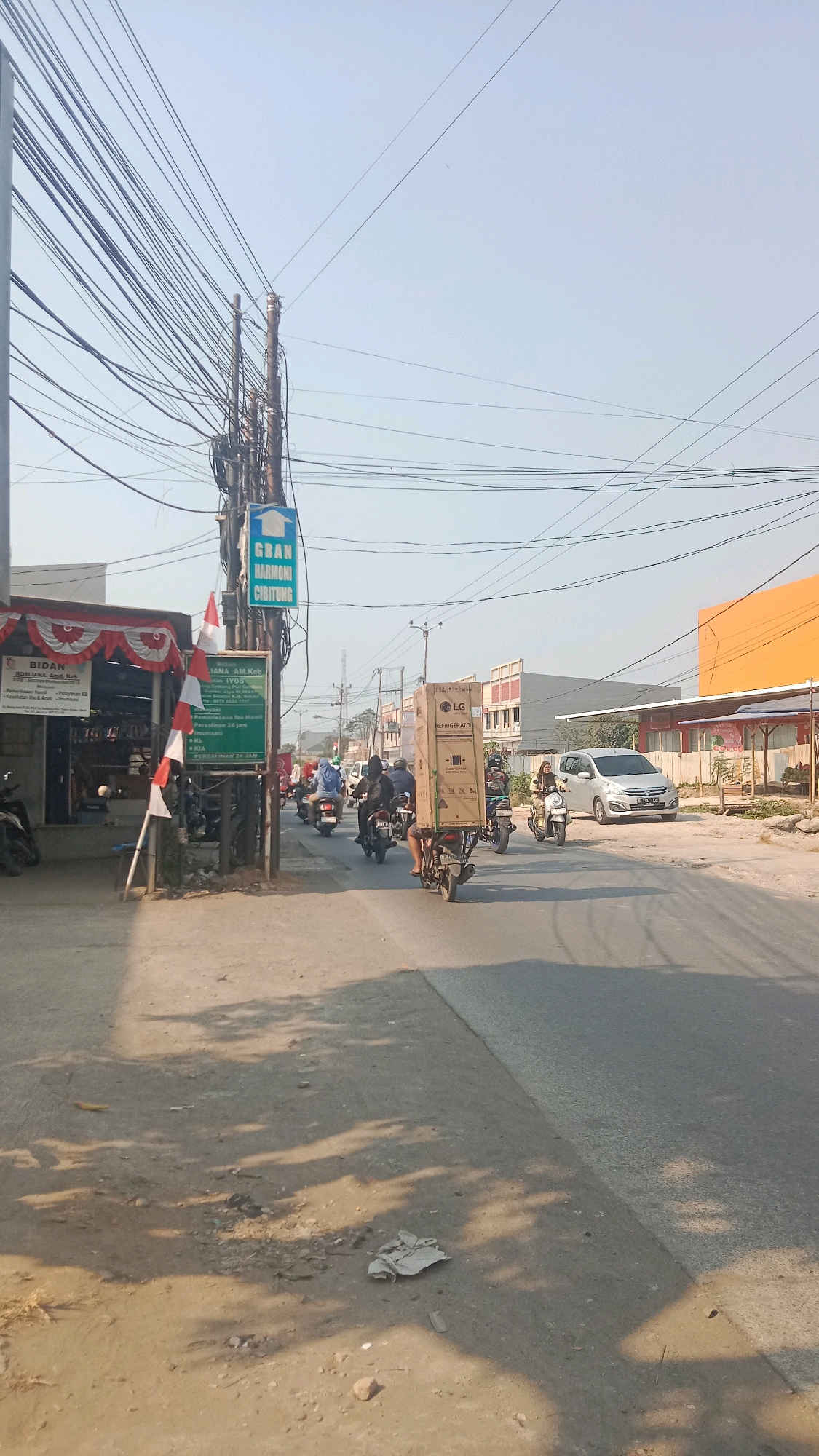 This is a street-level, eye-level, vertical photograph taken on a sunny day in Sumberjaya, Indonesia. The scene depicts a moderately busy street with several motorcycles and one car. The road is paved and appears to be in fair condition, with some debris and dirt on the shoulder. Overhead, a dense network of electrical wires is strung between utility poles, casting shadows on the road.
On the left side of the road, there is a building with a sign that reads "BIDAN ROSLIANA AMd. KEB." and another sign for "IJANA AM Keb." which appears to be a medical clinic. Red and white Indonesian flags are displayed on poles in front of these buildings.
Several people are visible, mostly riding motorcycles. One person on a motorcycle in the foreground is carrying a large cardboard box labeled "LG REFRIGERATOR" on the back. Other motorcycles are carrying passengers or goods. The car is a silver sedan, and it is moving away from the camera.
The background shows more buildings, some with orange and gray facades, and more utility poles and wires. The sky is clear and pale blue, indicating a bright day. The overall impression is a typical street scene in a populated area of Indonesia, with active commerce and transportation.