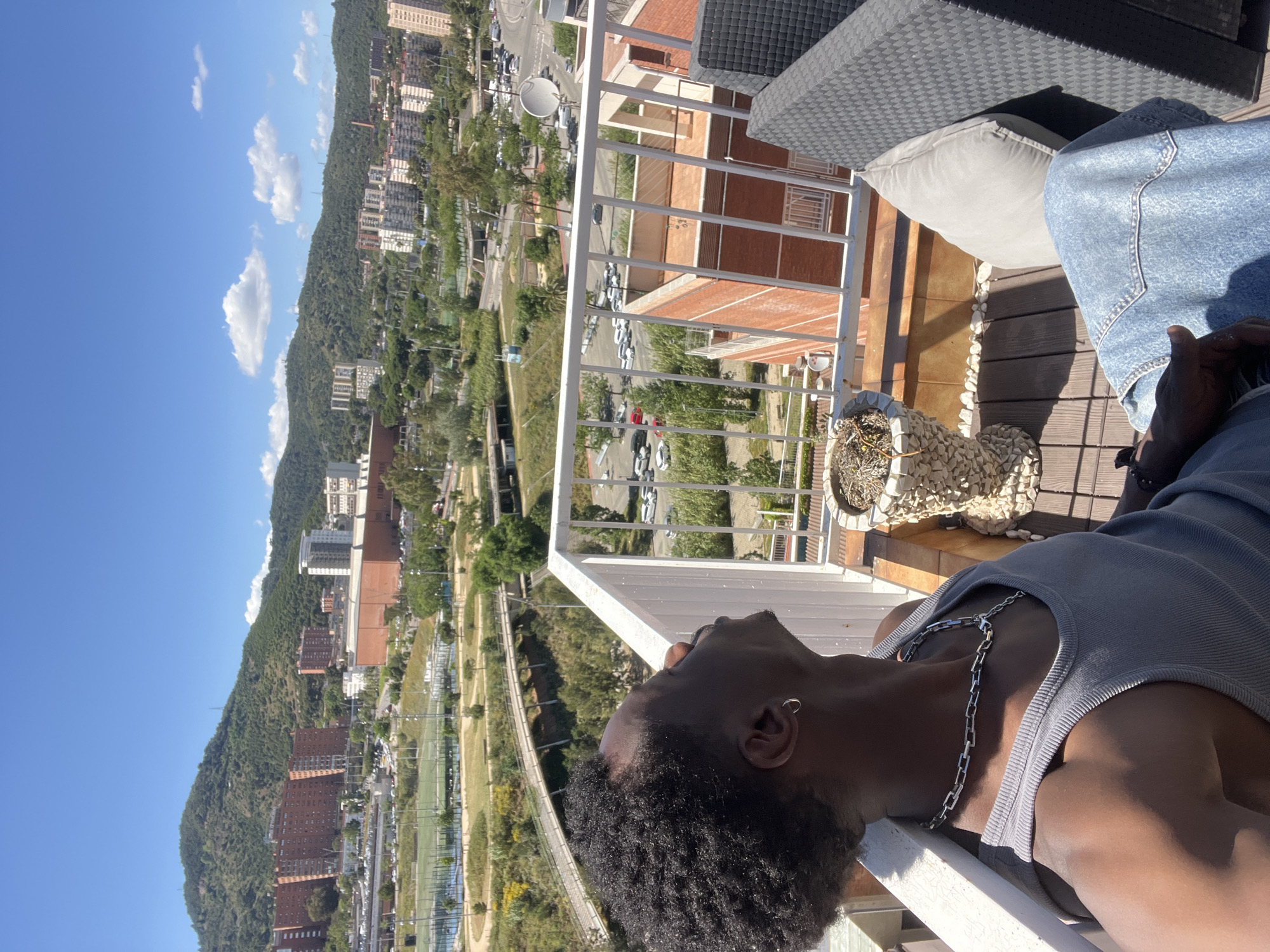A low-angle, horizontally oriented photograph captures a person reclining on a balcony with an expansive cityscape backdrop. The individual, who appears to be a young man, has dark skin, short curly hair, and is wearing a gray tank top with a silver chain necklace. He's positioned leaning against the white railing of the balcony, with his head tilted back looking upward, away from the camera. His right ear, adorned with a small hoop earring, is visible.
The balcony features brown wooden decking, a gray wicker chair with a gray cushion, and a large decorative vase filled with dried flowers or small pebbles. Portions of denim fabric suggest another individual is also present on the balcony, though not entirely visible.
Beyond the balcony railing, a densely populated urban landscape unfolds. Buildings of varying heights and architectural styles, predominantly in shades of red brick and concrete, populate the scene. A winding road, presumably a highway, snakes through the landscape. Trees are interspersed, and a lush, green hill dominates the background. The sky is a clear, bright blue, dotted with a few puffy white clouds.
While the exact location isn't explicitly mentioned, the distinctive urban landscape bears a resemblance to the city of Bilbao, Spain. However, the prompt is incorrectly asking for a description of Barcelona, Spain.