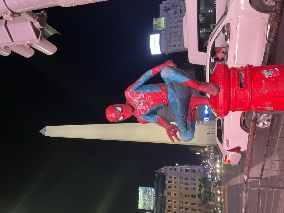 The image displays a nighttime urban scene in Buenos Aires, Argentina, at the Plaza de la República, with the Obelisco de Buenos Aires prominent in the background. A person dressed in a red and blue Spider-Man costume, featuring a spider emblem and web pattern, is crouched atop a red cylindrical public mailbox in the lower right foreground. The costumed figure's left hand is extended forward, with the index and middle fingers raised. In the mid-ground, a white pickup truck, with a male driver and female passenger visible inside, is stopped or moving slowly on a city street. Other vehicles and a metal railing are also present. The Obelisco is brightly illuminated against a dark sky, and various city buildings with lighted windows and advertising billboards (one reading "Sarmiento") line the streets. A purple-hued traffic light fixture is partially visible in the upper right.