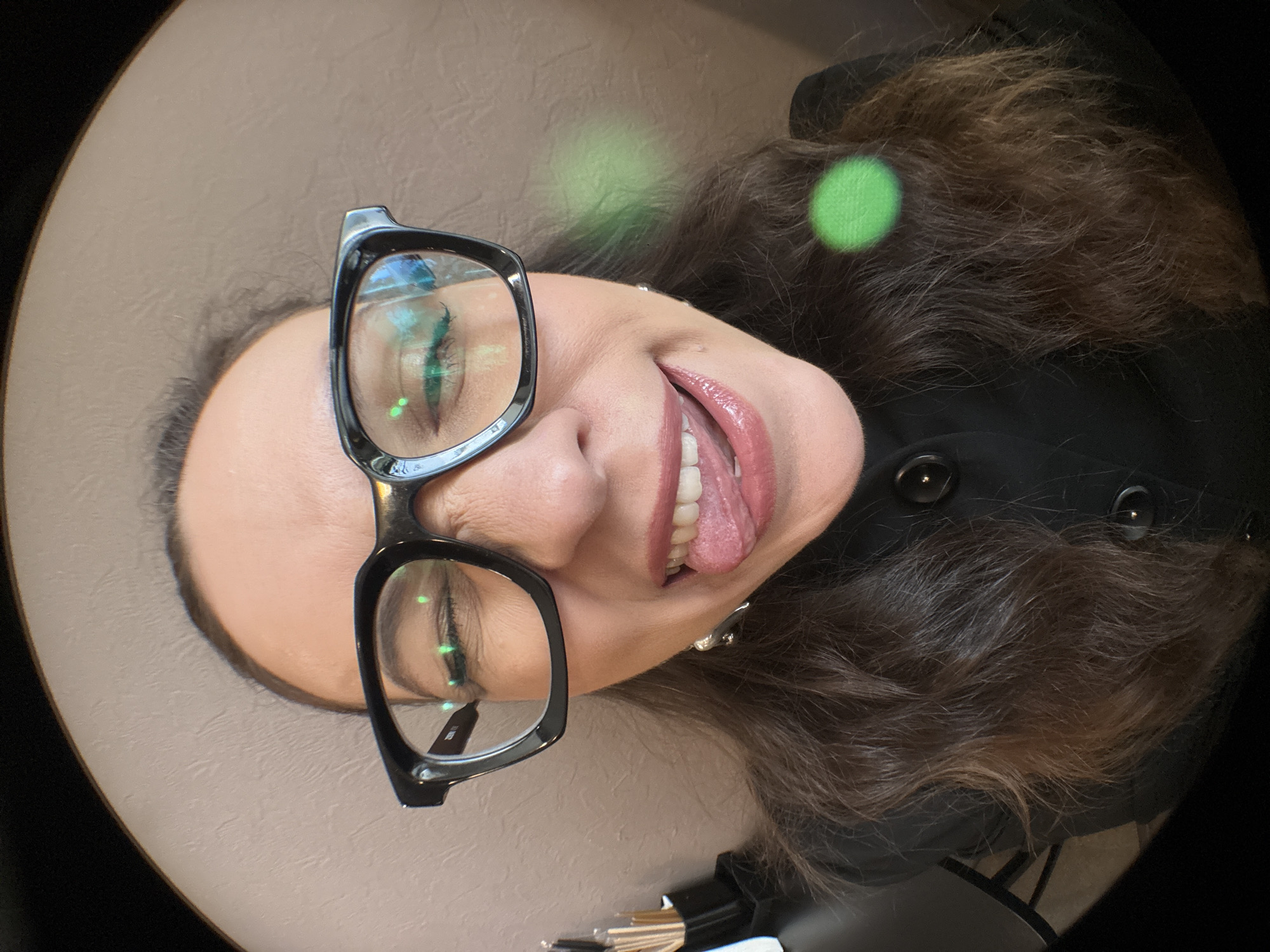 A fisheye lens close-up portrait shows a woman with dark brown hair wearing black-rimmed glasses and a black collared coat. She is sticking her tongue out with her eyes closed, and her lips are painted with a glossy lipstick. There are two green light refractions visible in the image, one on her hair and one to her right. The background appears to be a textured, light brown wall. The scene is indoors, but no specific location in the city of Chaikovsky, Russia can be determined from the visual information.