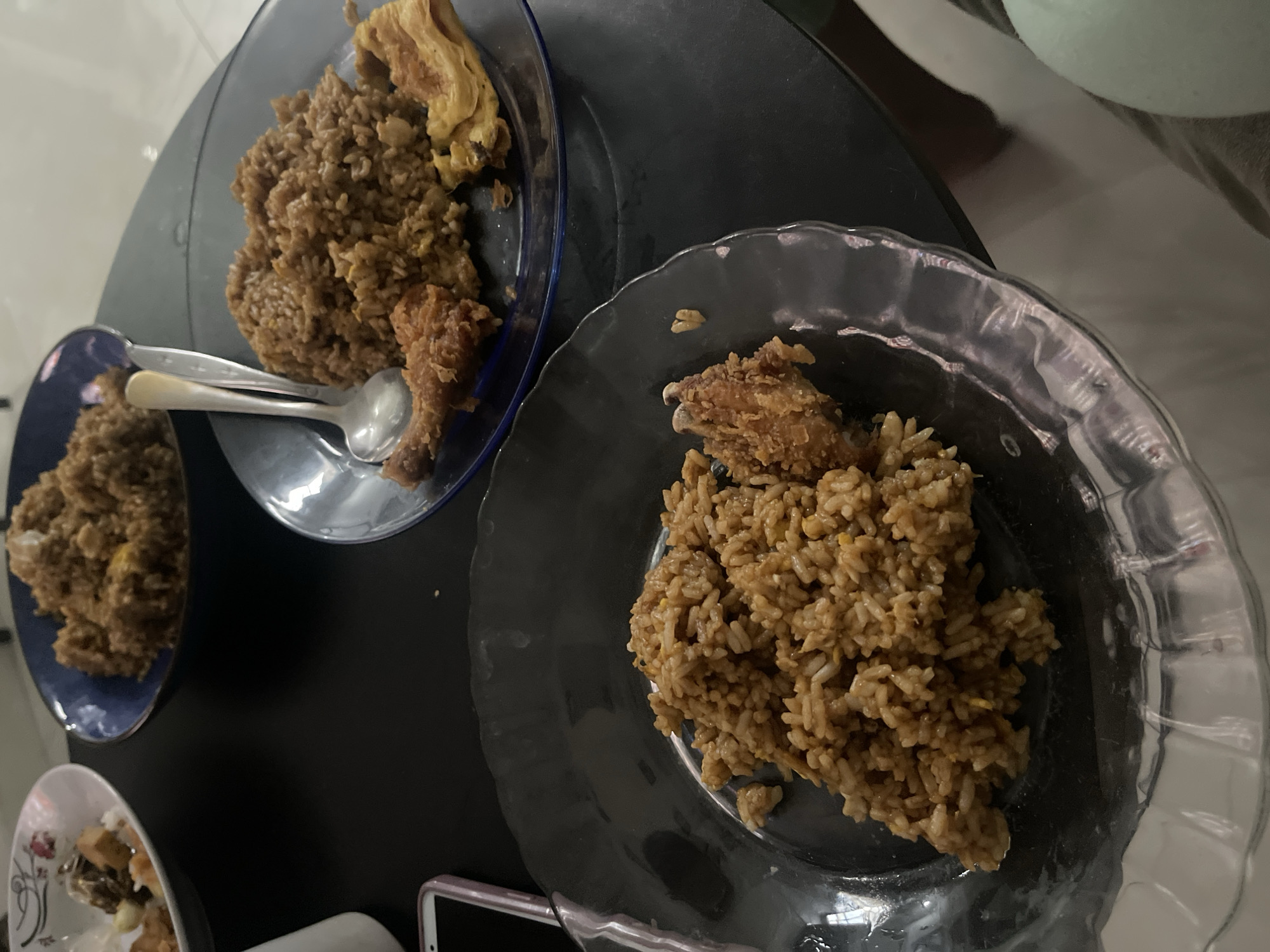 The image shows a close-up view of several plates of food on a dark, reflective surface, likely a table. The main subjects are plates filled with fried rice and fried chicken.
In the upper left of the image, a dark blue plate holds a serving of fried rice with pieces of fried chicken and what appears to be an egg omelet. Two spoons are resting on the plate. To the left of this plate, another smaller blue plate is partially visible, also containing fried rice.
In the foreground and to the right, a clear, fluted glass plate is filled with fried rice and a piece of fried chicken. Another small bowl with food is visible in the lower left corner. The overall scene suggests a meal is being served or has been prepared. The lighting is artificial, and there are no obvious cues about the time of day or weather. The setting appears to be indoors, possibly a dining area. No text is visible in the image.