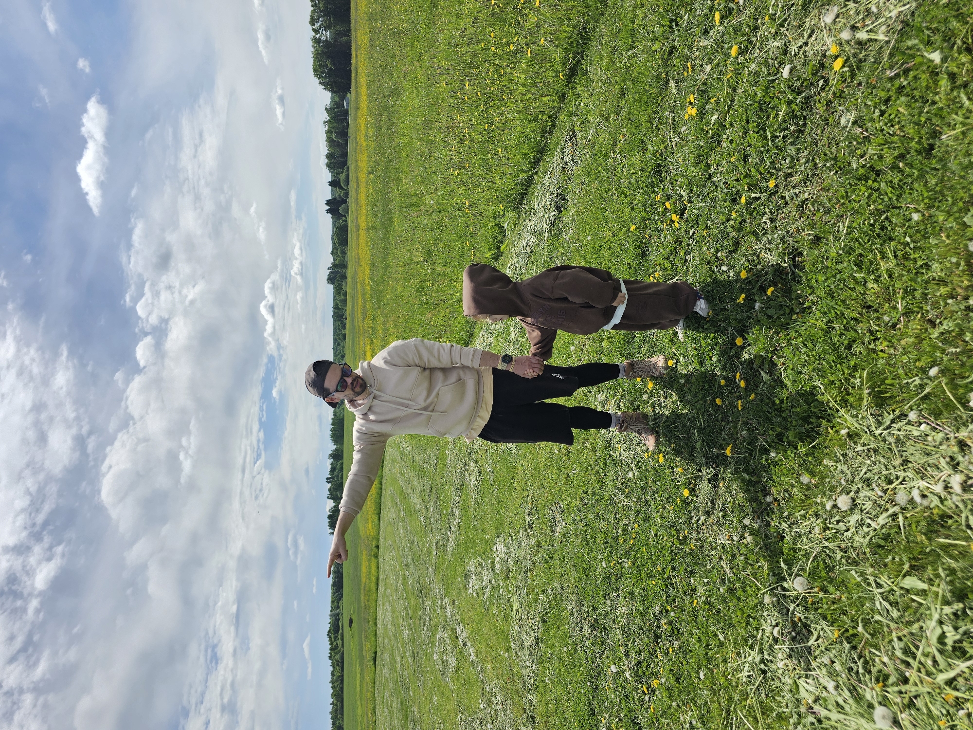 A medium shot depicts a man and a child standing in a field. The man, wearing a beige hoodie, black shorts, brown boots, sunglasses, and a hat, is holding the hand of the child. The child is wearing a brown hooded sweatsuit and white tennis shoes. The man is pointing toward the left side of the frame with his other hand. The field consists of grass and dandelions. The sky is visible in the background and contains blue sky and white clouds. A tree line appears on the horizon. The location is Unknown, Unknown.