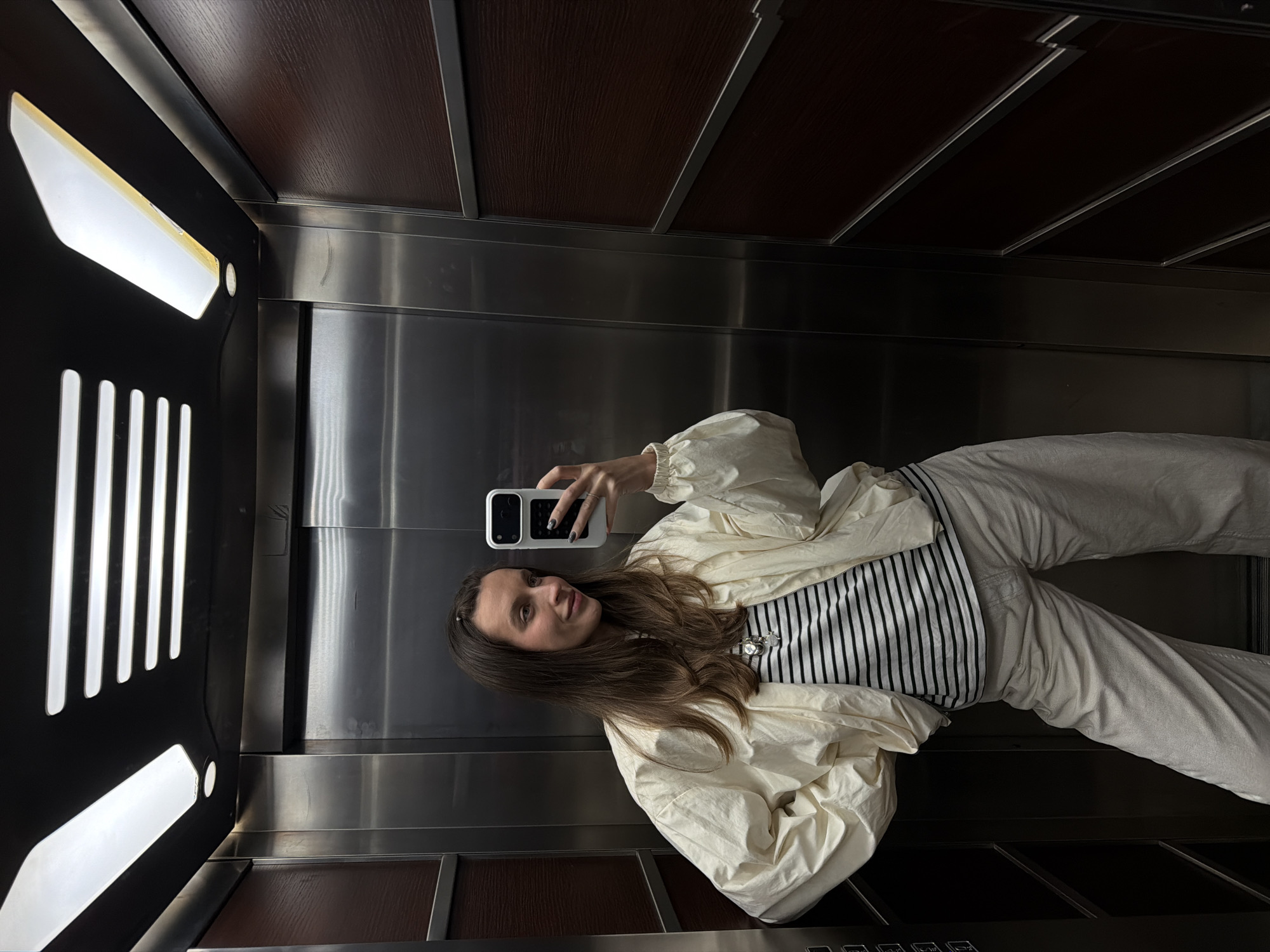 The image shows a young woman in an elevator, taking a selfie with her smartphone. She is wearing a white jacket, a black and white striped top, and light-colored pants. The elevator interior features a stainless steel wall panel, a dark wood paneling on the top and bottom, and a black control panel with white text. The lighting is provided by elongated white lights integrated into the black paneling. There is no indication of the location being in Budva, Montenegro, or any city for that matter.