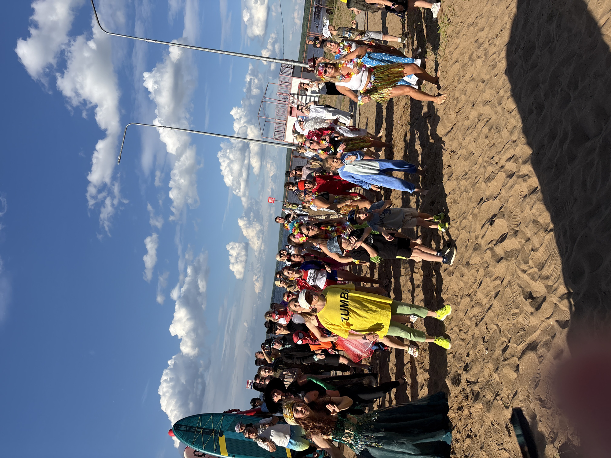The image depicts a group of people gathered on a sandy beach under a bright blue sky with scattered white clouds. They appear to be participating in an outdoor event, possibly a costume party or themed gathering, given the variety of attire. Many individuals are wearing leis, hula skirts, and colorful outfits. One person in the foreground wears a yellow shirt with the text "ZUMBA" in black letters. In the background, there's a sandy area, some low-lying vegetation, and what looks like a structure with metal poles and railings. Two tall, thin metal poles with lights are visible in the sky, likely part of the beach infrastructure. The sun appears to be high in the sky, suggesting it is daytime, and the weather seems pleasant. The overall mood seems festive and relaxed. Two paddleboards are also visible in the lower part of the image. The scene is set on a beach, with a body of water likely present beyond the sandy area.