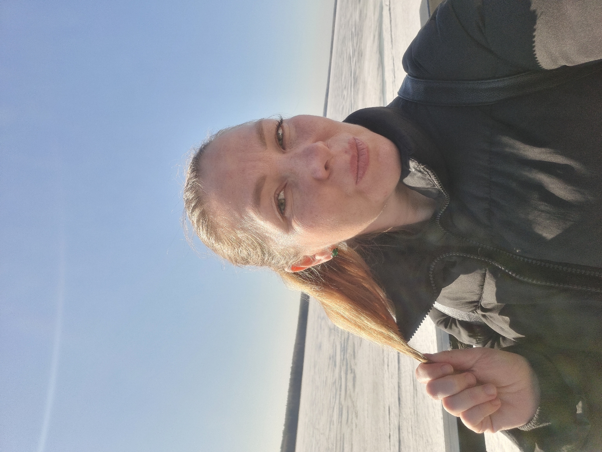 A close-up, brightly lit selfie features a woman outdoors on a clear, sunny day. The woman, with fair skin and light-colored hair pulled back, looks directly at the camera with a neutral expression and slightly parted lips. Her eyes are light-colored, and a small green stud earring is visible in her right ear. She is dressed in a dark, possibly black, winter jacket with a visible zipper and textured fabric. Her right hand is shown in the lower right portion of the frame, gently holding a strand of her ponytail.
The background is divided between a vast, clear blue sky occupying the left side of the frame and a wide expanse of snow or ice covering the ground on the right, stretching into the distance. A faint, dark line, likely distant trees or land, can be seen where the horizon meets the sky. The bright sunlight suggests it is daytime, and the winter attire and snowy landscape indicate a cold environment. The image evokes a tranquil, crisp winter day in what is contextually known as Chaikovsky, Russia. There is no visible text in the media file.