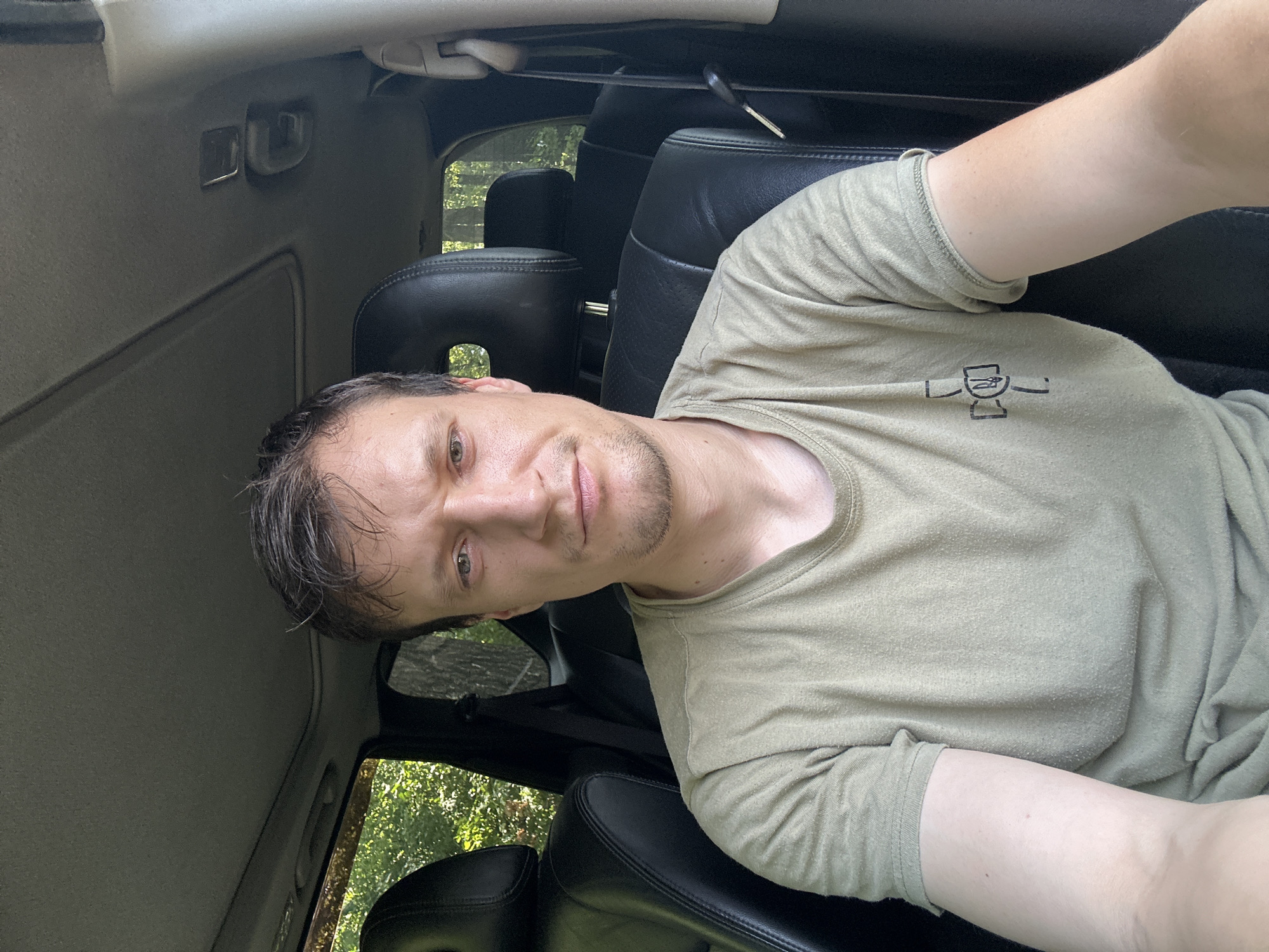 In the driver's seat of a car, a Caucasian male with short brown hair and light skin is captured in a selfie. He is wearing a light olive-green t-shirt with a black graphic design on the left chest. The man has a neutral expression, looking directly at the camera. The interior of the car is visible, with black leather seats and a sunroof. The exterior view through the windows shows a leafy, green environment suggesting a location with trees. The image appears to be taken outdoors, likely on a sunny day, given the lighting. The scene does not provide any specific indicators to place it within the city of Makiv, Ukraine.