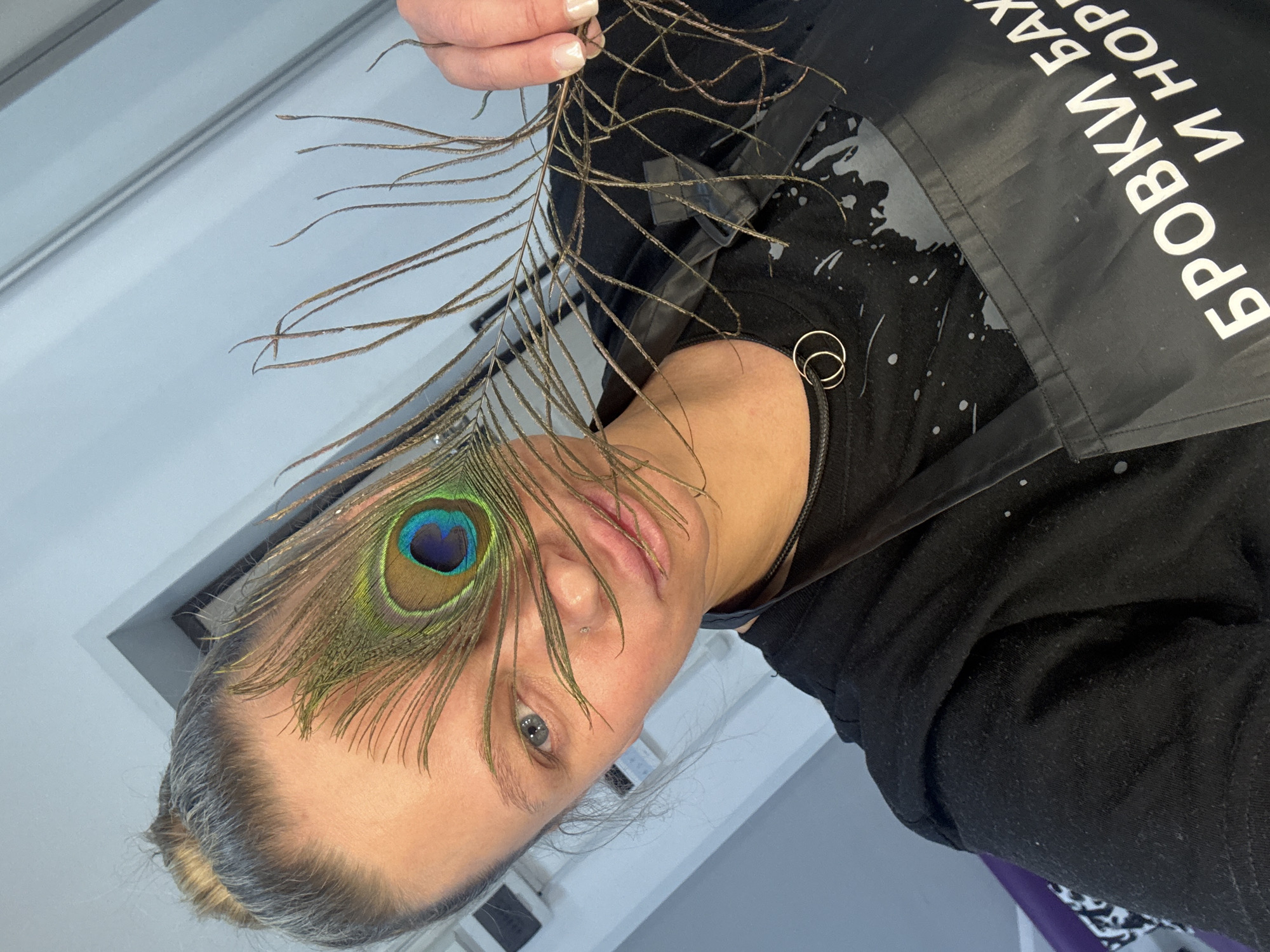 A person is holding a peacock feather over their left eye. The person has gray hair, light skin, and is wearing a black shirt and a black apron with white text in Cyrillic, possibly in the city of Perm, Russia. A window frame is visible in the background. The person's fingers are visible holding the stem of the feather. They appear to have a nose ring on the left side of their nose. There are two small gold rings around a black cord on their shoulder. The apron has splotches of grey and white paint.
