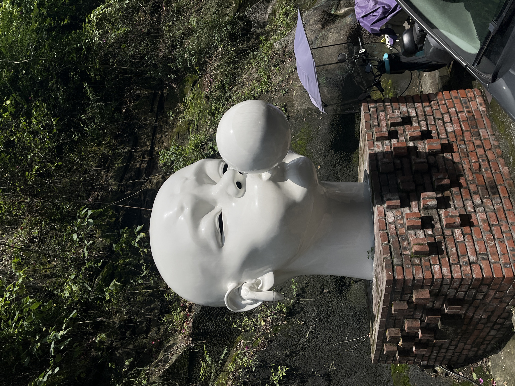 The image depicts a large, white, stylized sculpture of a child's head on a brick pedestal, set against a lush, green, overgrown hillside. The sculpture's mouth is open, with a large, white bubblegum bubble blowing from it. The child has closed eyes and is wearing what appears to be a white earbud in its left ear. The brick pedestal is constructed with a decorative pattern of protruding bricks. To the right of the sculpture, a scooter is visible, partially covered by a purple tarp. The background consists of a steep, rocky slope covered in dense vegetation and trees, suggesting an outdoor, possibly rural or park-like setting. The lighting indicates it is daytime with sunlight casting shadows. While the location is stated to be the Chongqing High-tech Zone, China, the visual elements themselves do not provide specific indicators of this precise location beyond its general characteristics. There are no people visible in the image.