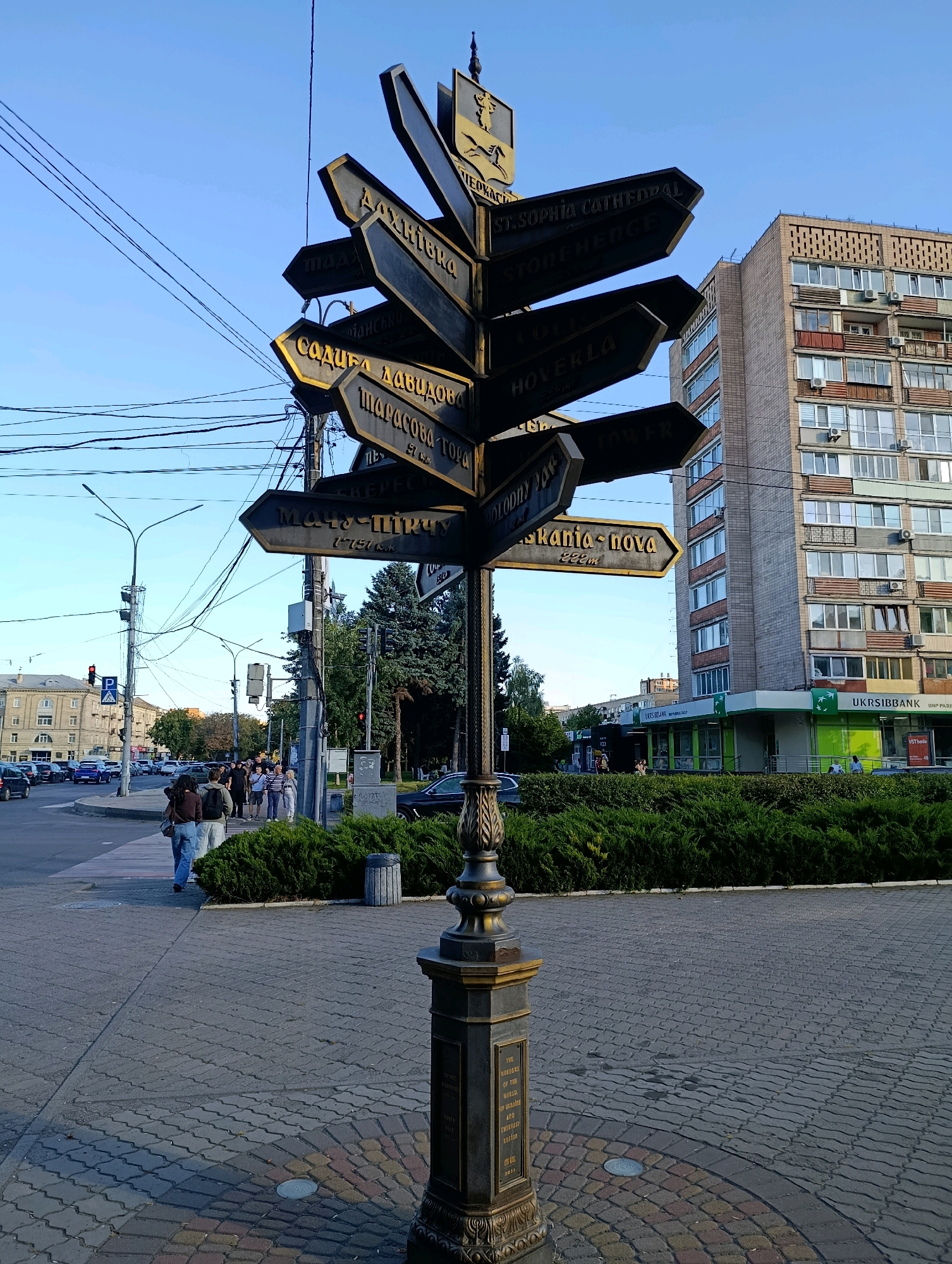 This image captures a street scene in Cherkasy, Ukraine, featuring a prominent directional signpost. The signpost, made of dark metal with gold lettering, displays multiple directional arrows pointing to various locations, some in English and others in Cyrillic script. One sign indicates "ST. SOPHIA CATHEDRAL" and another "HOVERLA 950m". The base of the signpost has an inscription that appears to be in English, including the text "THE BROTHERS OF THE WORLD OF UKRAINIAN AND CHERKASSY REGION". The scene is set on a paved sidewalk with a circular brick pattern at the base of the signpost. Across the street, a multi-story brick building with numerous windows and balconies dominates the background. A bank with green signage reading "UKRSIBBANK" is visible at street level. Traffic lights and street lamps are present, and several cars are on the road. A group of people are walking on the sidewalk near the signpost. The sky is clear and blue, suggesting daytime. The overall atmosphere is that of a typical urban environment in Ukraine.