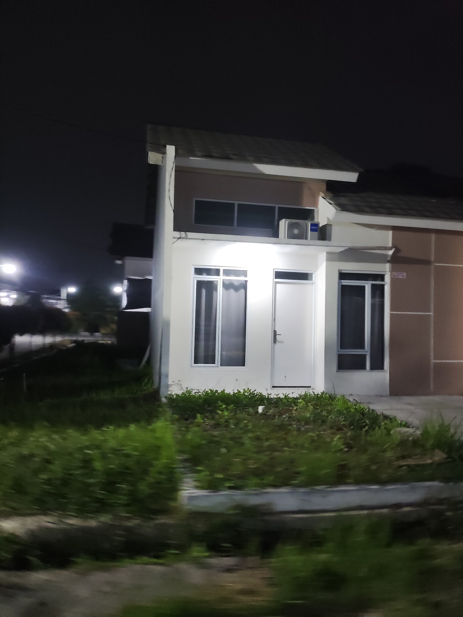 The image depicts a two-story house at night in the city of Wanajaya, Indonesia. The house has a light-colored exterior with dark-colored roofing. It features several windows with white frames and frosted glass, a white front door with a dark handle, and an air conditioning unit mounted on the upper portion of the exterior wall. The foreground is dominated by an unkempt grassy area with some dirt patches, suggesting a lack of recent landscaping. In the background, streetlights illuminate a blurred street and some trees. The overall atmosphere is dark and somewhat desolate.