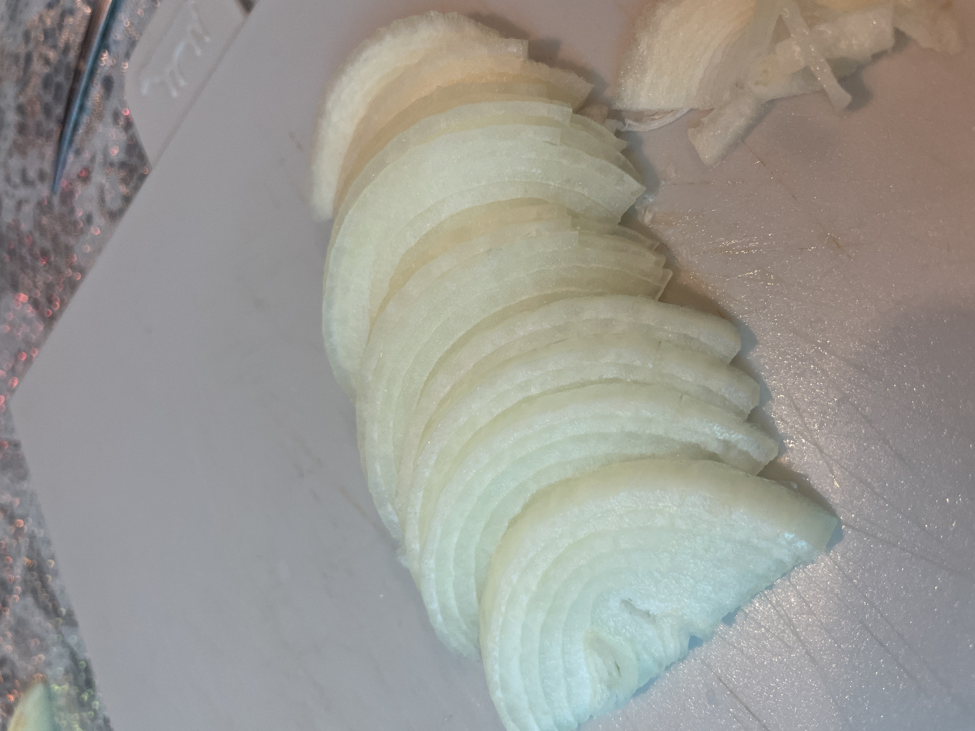 The image shows a pile of thinly sliced white onions on a white cutting board. The onions are stacked in a slight curve. Some loose onion pieces are scattered to the right of the stack. A small portion of a counter, possibly granite or a similar material, and what appears to be a kitchen utensil are visible in the upper left corner. The image does not provide information about the location being in Olginka, Russia.