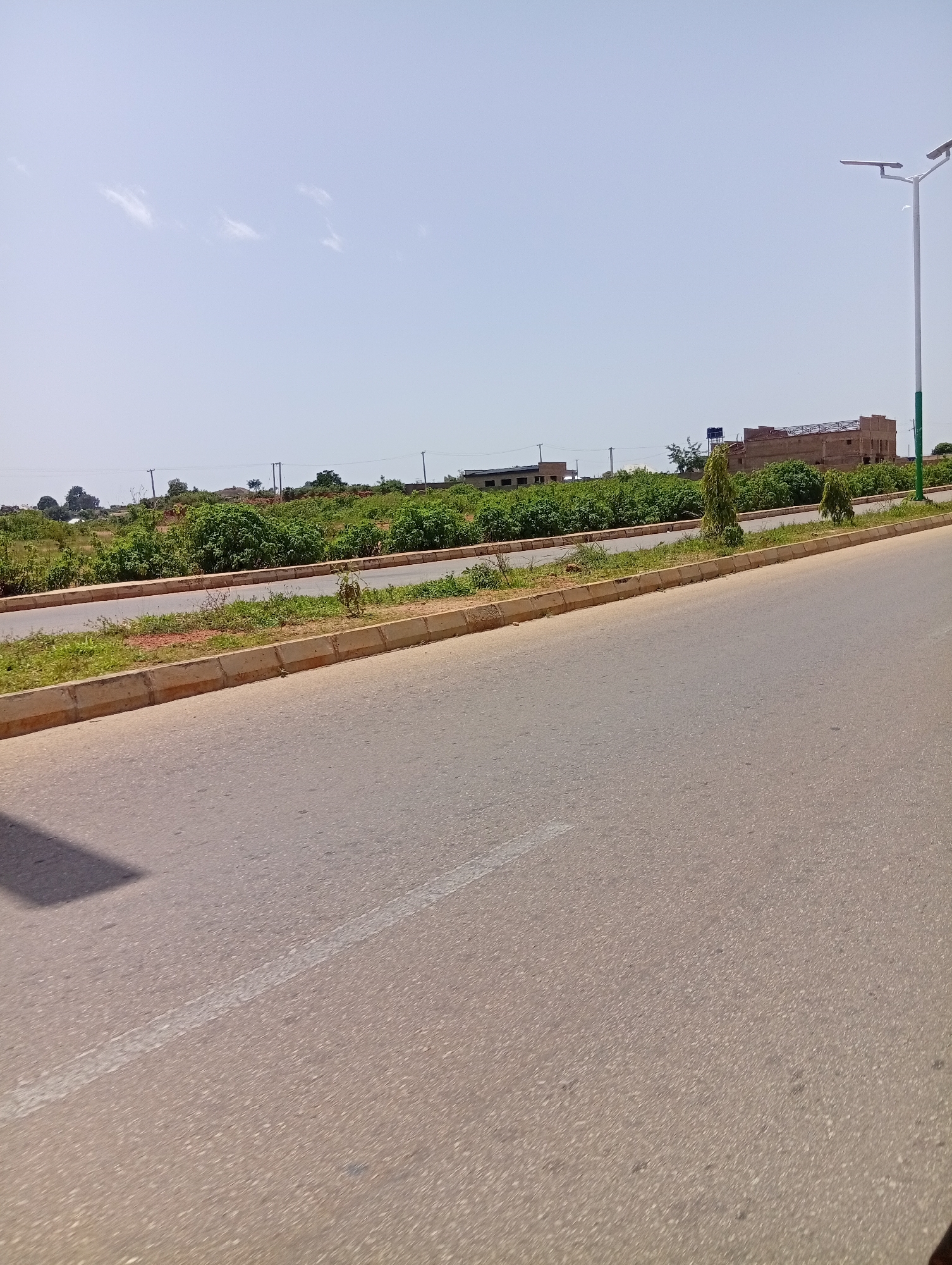 The image depicts a roadside scene in Jos, Nigeria. A paved road with a white dashed line runs diagonally across the frame from the lower left to the upper right. To the left of the road, a shadow is cast on the asphalt. The road is bordered by a raised concrete curb and a strip of grass. Beyond the grass, there is an area with sparse vegetation and some scattered debris. In the background, a dense line of green trees and bushes stretches horizontally across the midground. Further back, a large, unfinished building with a reddish-brown brick facade and exposed scaffolding is visible. A tall, white street light with two lamp fixtures stands to the right of the building. The sky is a pale blue with a few wispy white clouds. The overall environment appears to be an urban or semi-urban area undergoing development, characterized by infrastructure and natural elements.