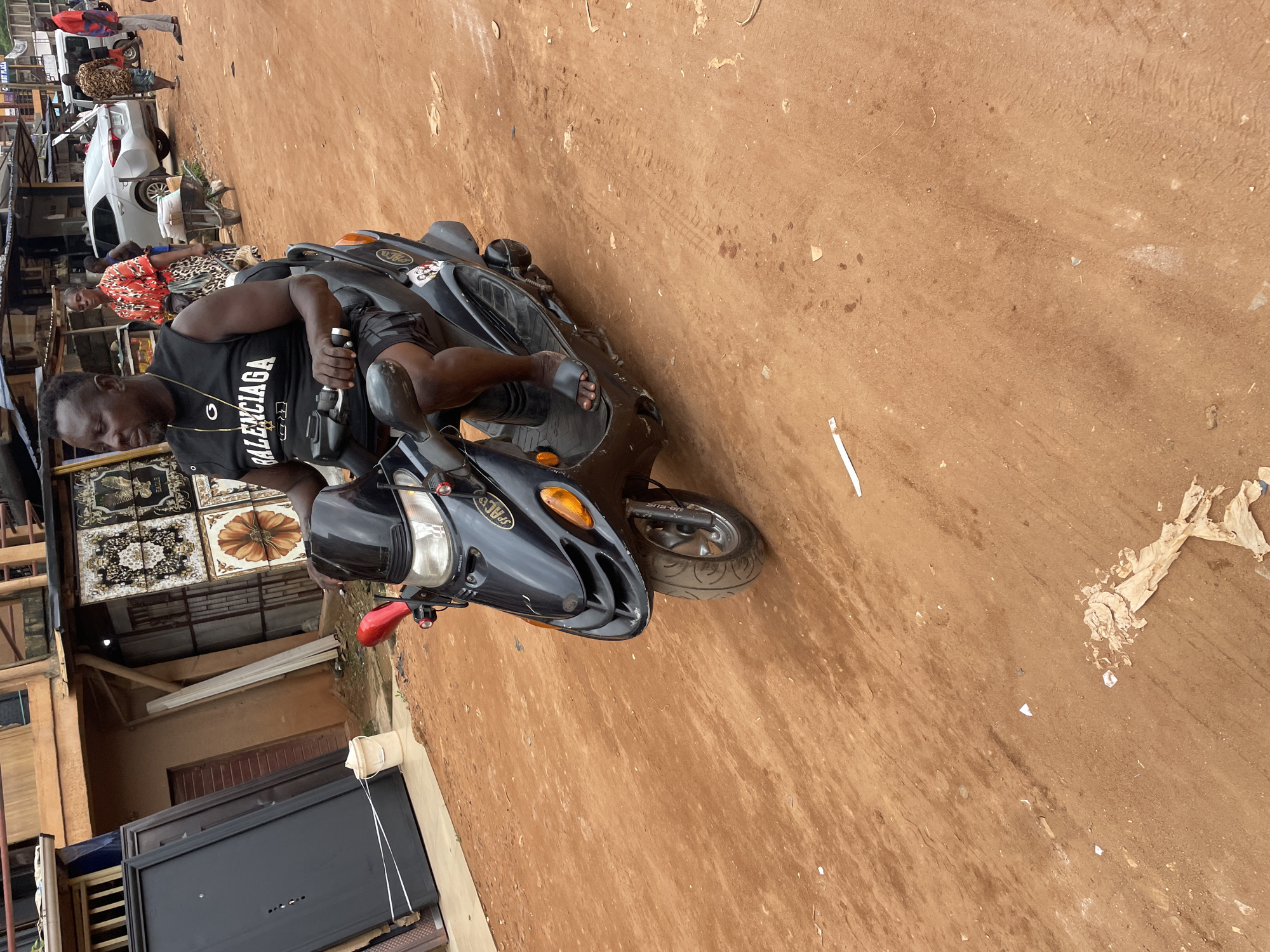 An outdoor, eye-level, medium shot shows a Black man riding a black scooter on a dirt road in Ogidi, Nigeria. He is wearing a black sleeveless shirt with a gold chain and necklace, black and white checkered shorts, and black sandals. The scooter is black with a red rearview mirror on the left side. The man has his hands on the handlebars and is looking to the left.
In the background, there are buildings with corrugated metal roofs, a silver car, and several other people. To the left of the scooter, there are large black doors leaning against a building. The ground is covered in reddish-brown dirt with some debris. The lighting suggests it is daytime.