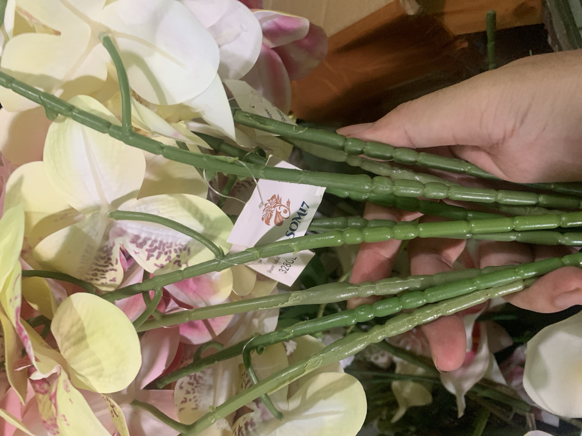 The image shows a close-up of artificial orchid stems being held by a person's hand. The stems are green and segmented, with some showing slight variations in color and texture. Behind the stems, blurred orchid flowers are visible in shades of yellow, pink, and white. A small white tag is attached to one of the stems, with some text and a logo. The background is out of focus and appears to be a brown surface, possibly a table or a container. There is no visible indication of the specific location being Kuala Pembuang, Indonesia, or any other city.